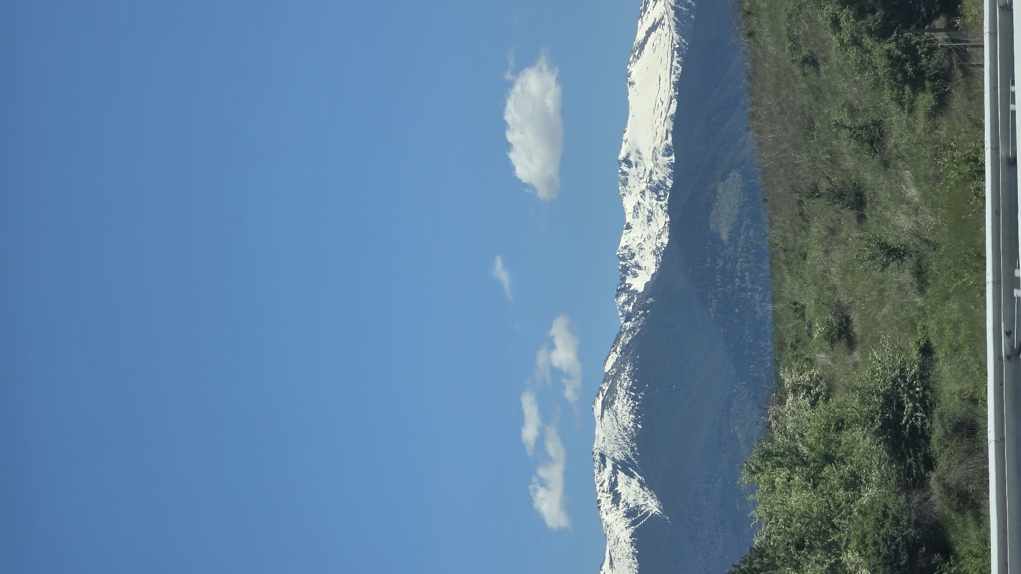 The image depicts a mountain range with snow-capped peaks against a bright blue sky with scattered clouds. To the right of the mountains, there is a green hillside. The picture also shows the metal structure of a bridge or railing along the right edge. There is no indication of the location being in Blatino, Bulgaria.