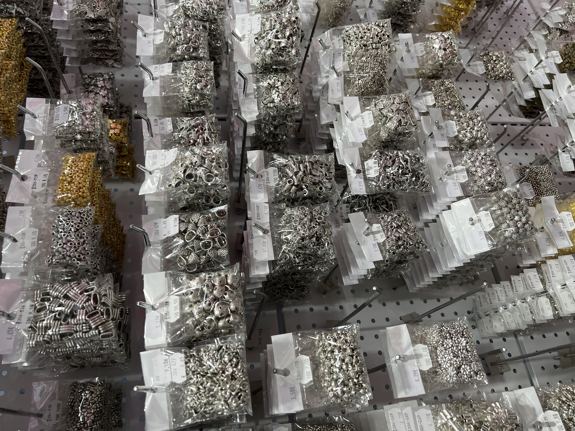 The image shows a display of various jewelry beads and components packaged in small clear plastic bags, hanging on metal pegboards. The beads are mostly silver-colored, with some gold-colored beads on the left side of the frame. Each bag has a white label with prices and identifying numbers printed in a European language (likely Bulgarian or related) using the Euro symbol (€). The display appears to be in a retail environment, possibly a craft or hobby store. The setting is a brightly lit commercial space with no people directly visible in the shot. No Burgas, Bulgaria, location has been identified.