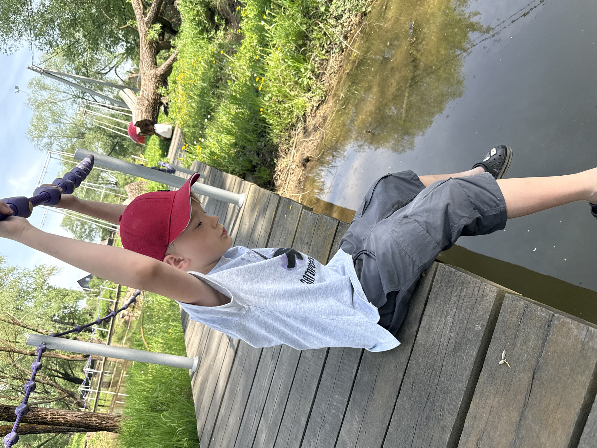 A child is hanging from a rope course over water. He wears a red baseball cap, a light gray T-shirt, and dark gray shorts, with black Crocs on his feet. He grips a purple rope ladder. The platform beneath him is made of wooden planks. The water reflects the green vegetation on the opposite bank. The location is Синьково, Russia.