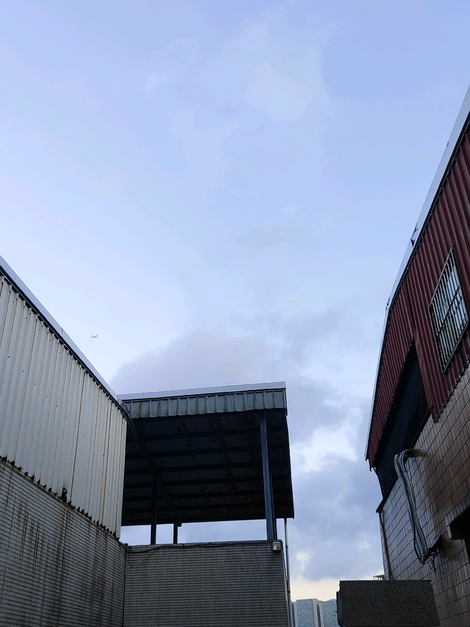 The image depicts an overcast sky above a cluster of buildings, likely industrial or commercial, in New Taipei City, Taiwan. Two structures are prominently featured in the foreground: one on the left with a ribbed, off-white metal facade and a brick-patterned lower wall, and another on the right with a corrugated, reddish-brown metal exterior and a brick facade. Between these two buildings is a structure with a dark, weathered metal roof supported by dark vertical beams, creating a covered area. The sky is a pale blue with scattered, wispy clouds. A single, small airplane is visible in the upper left portion of the frame. The overall scene suggests an urban, utilitarian environment.