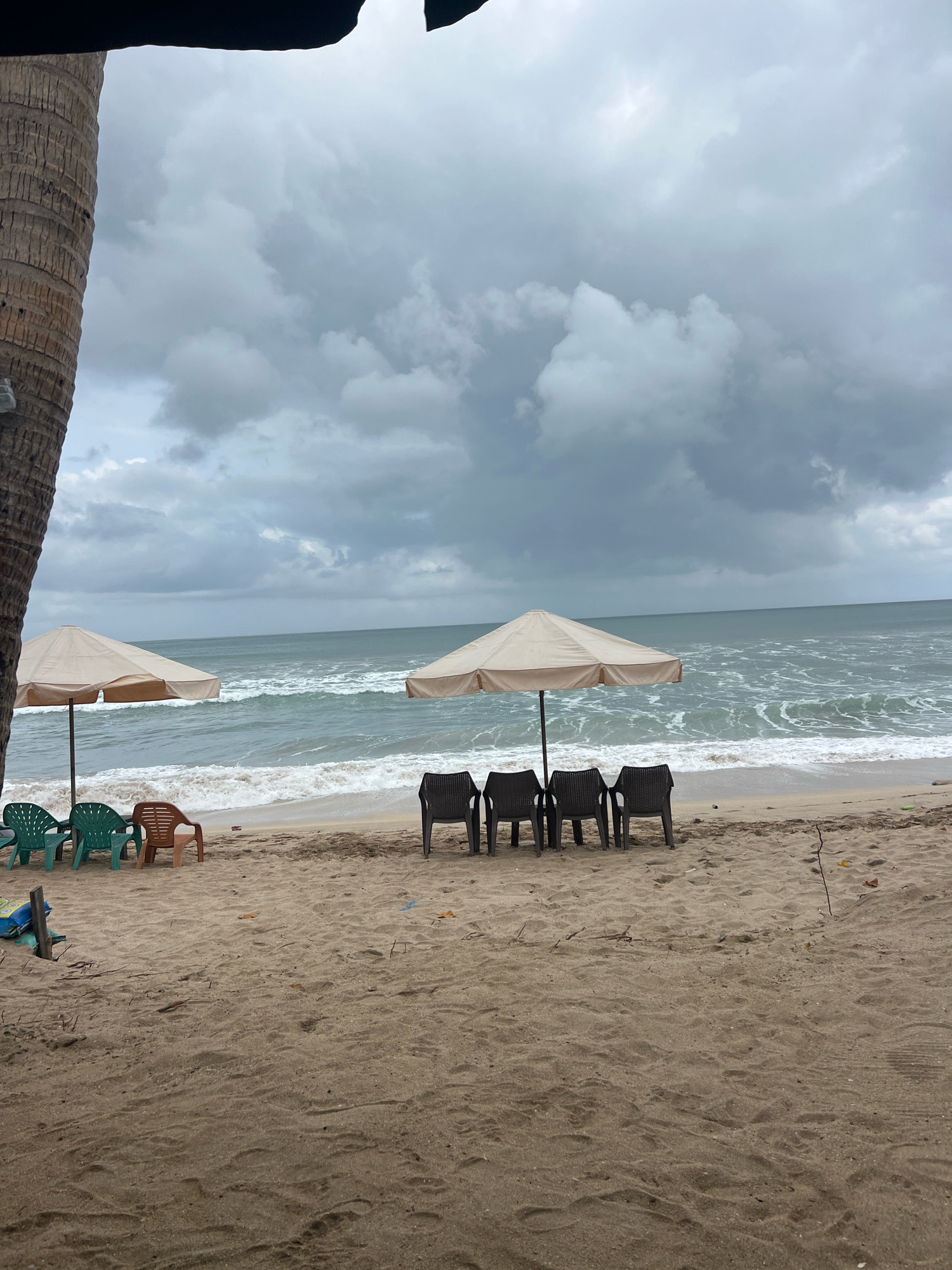 An overcast day at a beach in Kuta, Indonesia, with the ocean visible in the background. Two beige beach umbrellas are set up on the sand, each with chairs arranged in front of them. The umbrella on the left has three green plastic chairs and one brown plastic chair. The umbrella on the right has four black plastic chairs. A palm tree trunk is visible on the far left side of the image. The waves are breaking on the shore, and the sky is filled with dark, heavy clouds. The sand appears damp and somewhat uneven. There are no people visible in the scene.