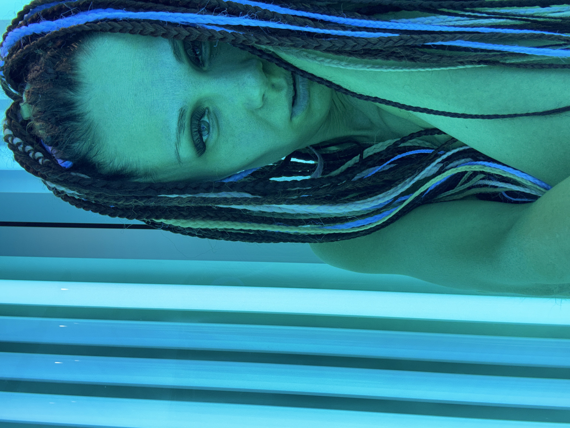 A person with braids lies in a tanning bed. The braids are a mix of dark brown, white, and blue colors. The lighting in the tanning bed gives the person a blue-green tint. The person's eyes are open and they are looking towards the camera. The tanning bed has a series of horizontal lights. The location is Unknown, Unknown.