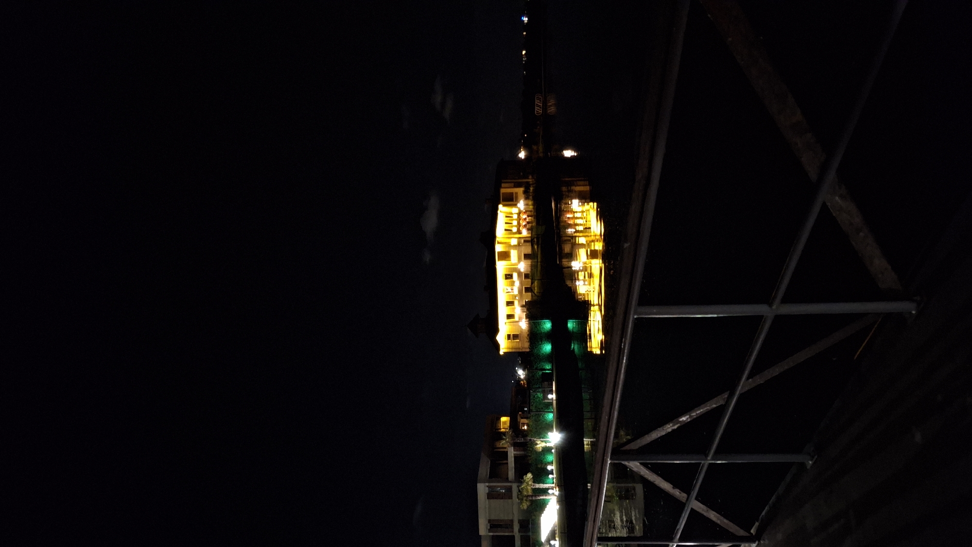 The image, taken at night, appears to show a building reflected in a body of water. The building is illuminated with yellow and green lights. Some kind of metal structure or railing is also in the foreground. The location is Казашко, Bulgaria.