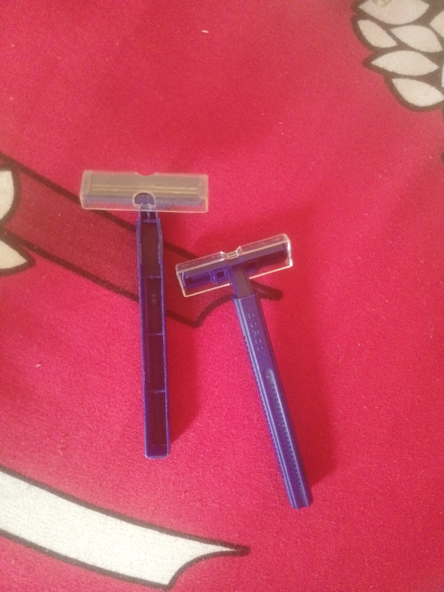 Two disposable blue razors with clear plastic protective covers are lying on a red textured surface. The razor on the left is oriented vertically, with its head pointing upwards. The razor on the right is angled to the upper right, with its head to the upper left. The red surface appears to be a cloth with a subtle pattern, possibly a rug or blanket, featuring cream-colored, stylized leaf-like shapes outlined in black. The image does not provide any information that could link it to Majemu, Nigeria or any specific city.
