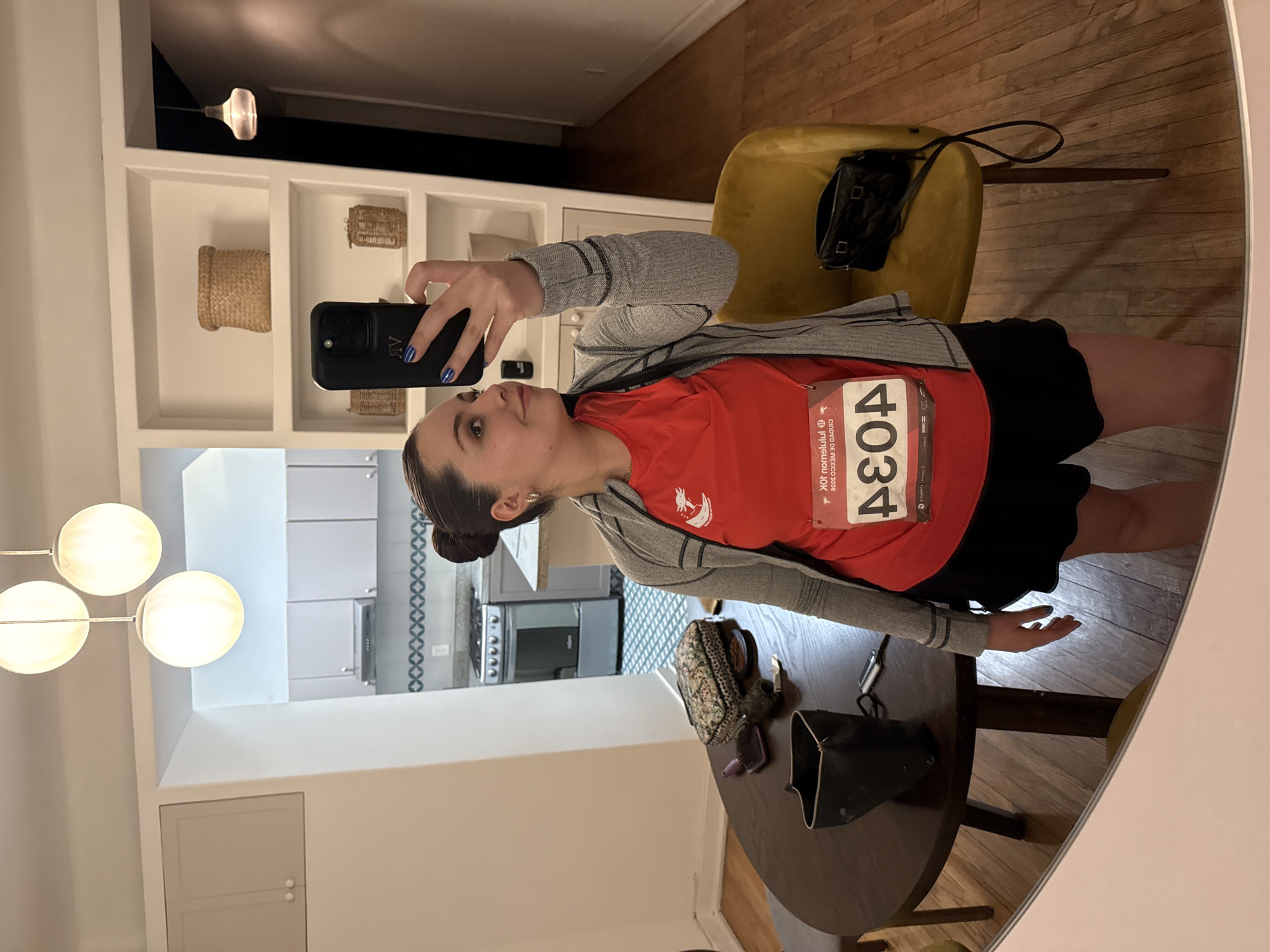 A woman is taking a selfie in a mirror, revealing a partial reflection of an interior space. She wears an orange t-shirt with a race number "4034" pinned to it, a gray jacket, and a black skirt. She holds a black cell phone with blue painted fingernails.
Behind her, the reflection shows a kitchen with light gray cabinets, stainless steel appliances, and a backsplash with a geometric pattern. Adjacent to the kitchen area is a dining space with a round dark wood table, various bags, and sunglasses. A yellow chair is visible, with a black purse placed on it.
A built-in white shelf unit with woven baskets is also seen. A multi-globe light fixture hangs nearby. The environment suggests a modern apartment setting, possibly located in Mexico City, based on the race bib's text.