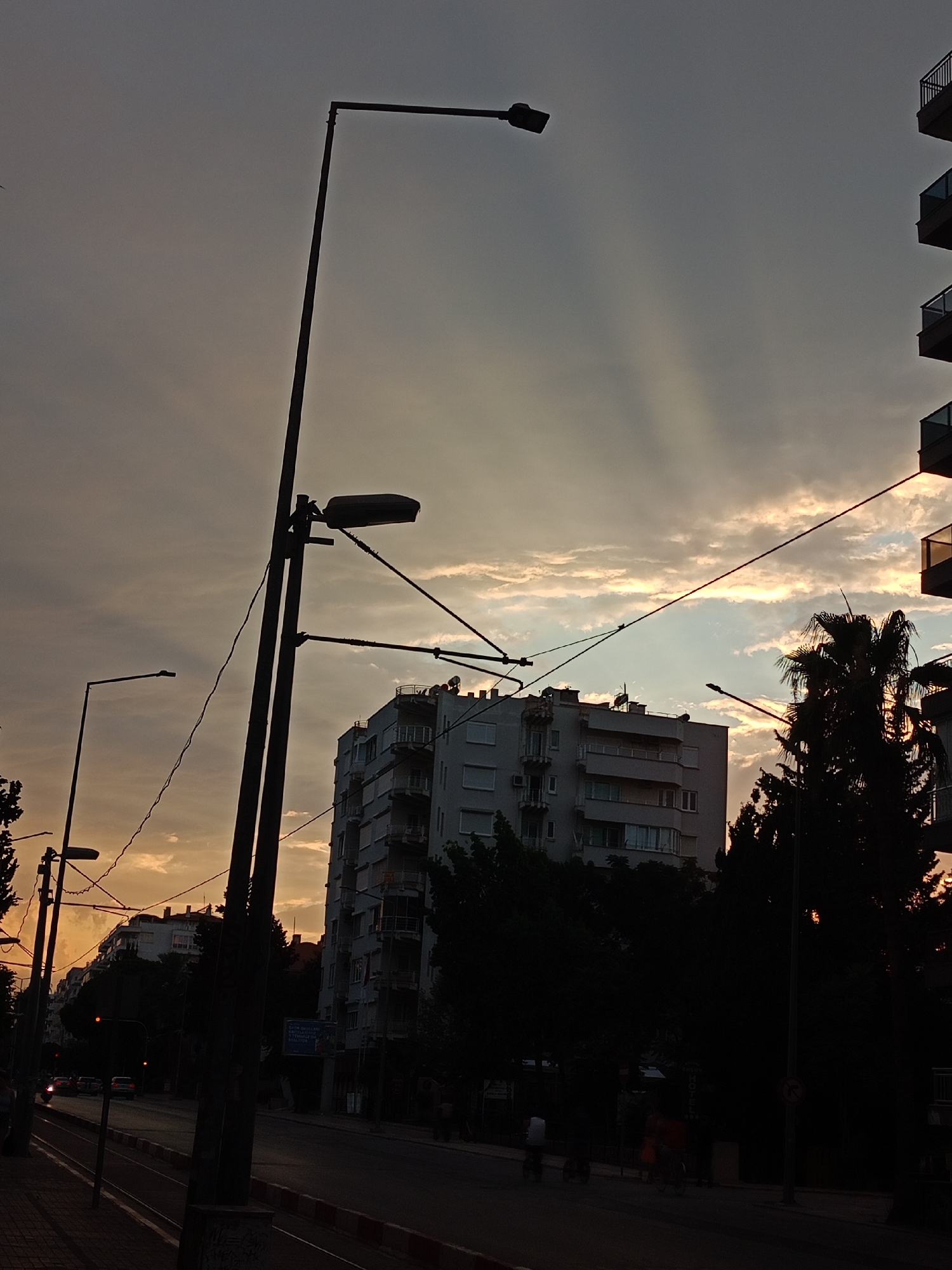 This is a vertical, eye-level, full shot of a street in Antalya, Turkey, at dusk. The sky is filled with crepuscular rays emanating from behind the clouds. In the foreground, silhouetted against the sky, are several tall streetlights and overhead electrical wires. To the right, a modern apartment building with balconies is partially visible. In the middle ground, silhouetted trees obscure much of the lower portion of the apartment building and the street. The street itself is dark, with a few vehicles visible, their headlights on. The sidewalk in the foreground is also dark. The overall impression is one of urban twilight, with the fading light casting long shadows and silhouetting the urban landscape.