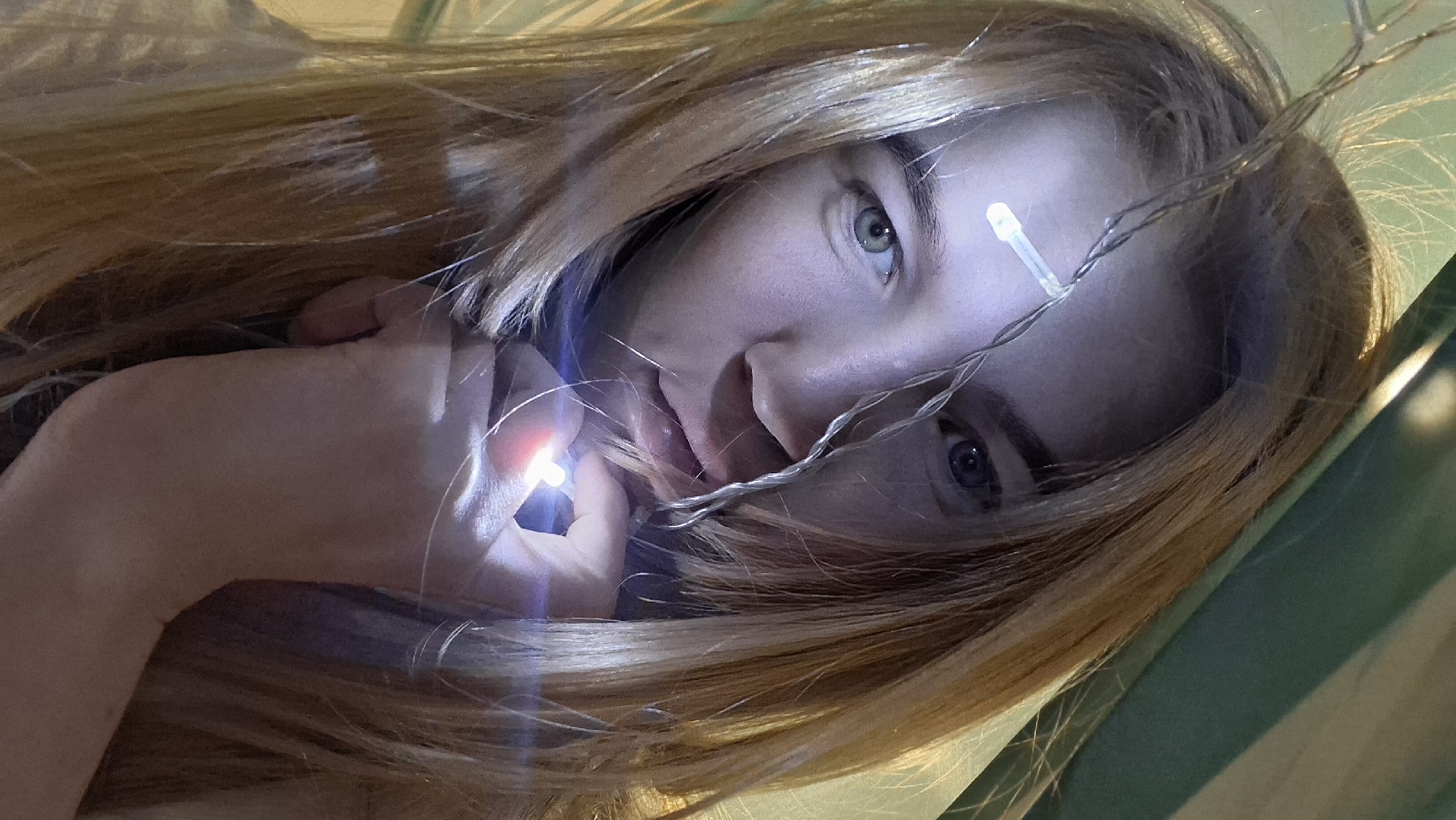 A fair-skinned woman with long, light brown hair is featured in this close-up shot. The image is angled, making the viewer look up at her. She has light-colored eyes and is holding a string of small LED lights, with one light near her forehead and another close to her mouth. The lights illuminate parts of her face and hair. The background has indistinct greenish tones. The location, Chaikovsky, Russia, cannot be inferred from the image.