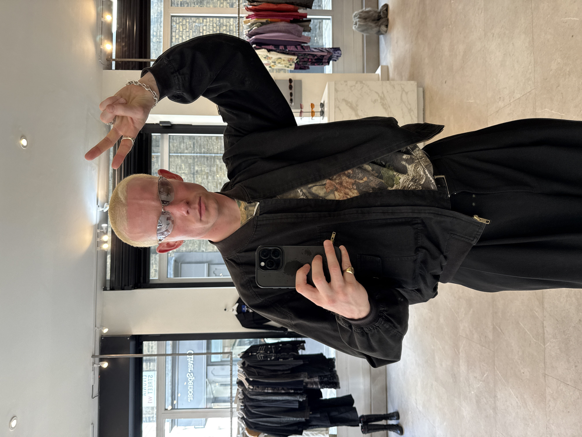 A young person with short, bleached blonde hair and reflective sunglasses is depicted taking a mirror selfie in what appears to be a clothing store. The image shows them from the torso up, facing the viewer. They are wearing a black zipped jacket over a camouflage-patterned t-shirt and black trousers.
With their left hand raised, the person is making a peace sign gesture, displaying a silver chain bracelet and several silver rings. Their right hand holds a black smartphone with a prominent camera array, capturing the reflection. A gold signet ring is visible on their right ring finger.
The background reveals the interior of a retail space. To the left, a light-colored wall with recessed lighting is visible. Behind the person, clothing racks hold various garments, including brightly colored tops and darker items. A display stand with sunglasses is seen on a white, marble-patterned surface. Through large windows in the background, a street view is partially visible, with reflections containing reversed text. Decipherable text includes "livre Spencer" and "STREET WI 33", suggesting an "Oliver Spencer" store, likely located on a street in London, United Kingdom. The flooring is a light-colored, possibly tiled or stone, surface. The overall lighting suggests it is daytime.