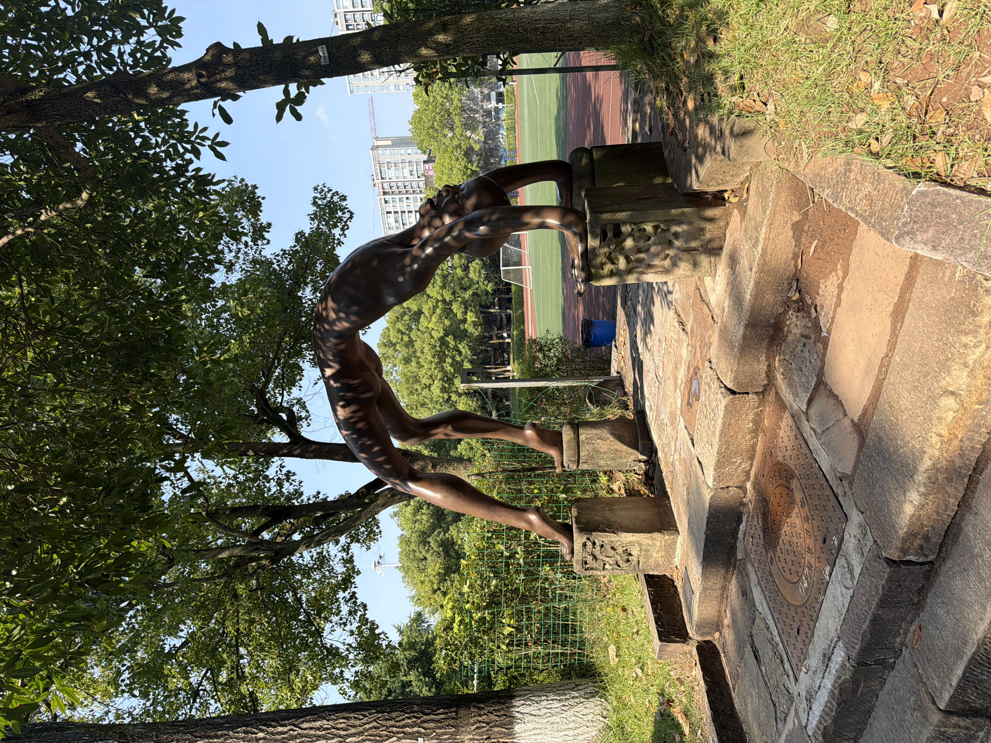 A bronze statue of a person in a backbend pose is situated on stone pedestals in an outdoor park setting. The statue is positioned on a paved walkway with stone steps leading up to it. The background features lush green trees, a grassy area, and a sports field with a running track and soccer goals. In the distance, modern high-rise buildings and a construction crane are visible under a clear blue sky. The lighting suggests it is daytime, with shadows cast by the trees and statue. The location is identified as the Chongqing High-tech Zone, China.