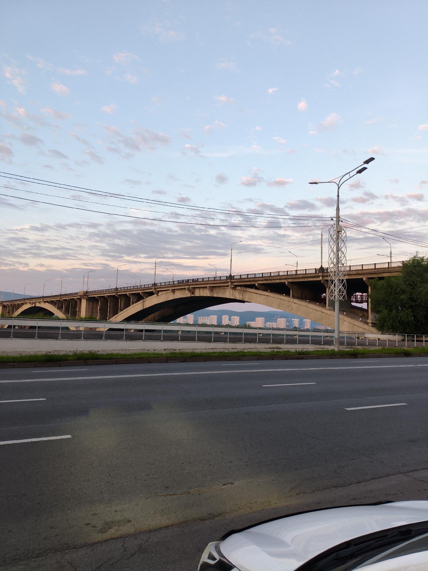 The image captures a wide, concrete bridge spanning a body of water, likely the Yenisei River in Krasnoyarsk, Russia. The bridge features multiple arches and is illuminated by streetlights, casting long shadows. A highway runs in the foreground, with lane markings clearly visible. In the distance, a cluster of tall buildings forms a city skyline. The sky above is a pale blue, dotted with wispy pink and white clouds, suggesting either sunrise or sunset. A street lamp with two lights stands on the right side of the frame. The overall scene depicts an urban landscape at dusk or dawn.