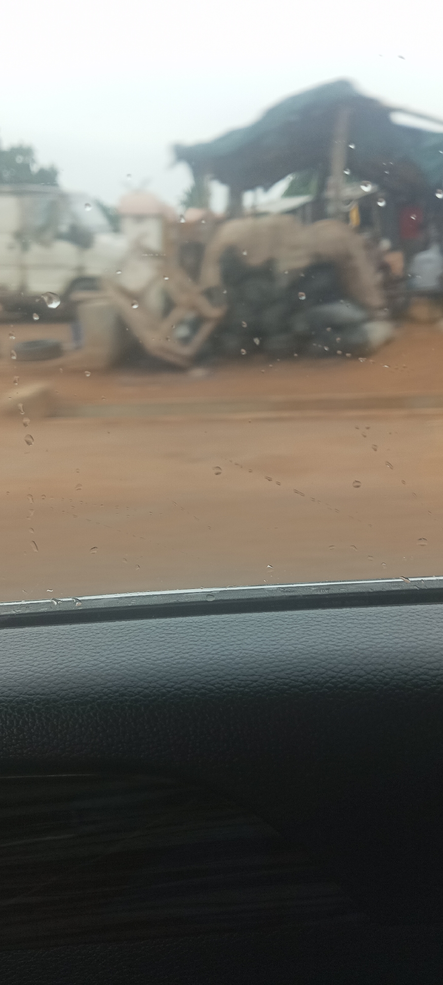 The image, taken from inside a vehicle on a rainy day in Gwadanye, Nigeria, shows a blurred view of a roadside scene. Through the rain-streaked window, a red-brown dirt road is visible. On the side of the road, there's a parked white vehicle. A temporary structure, possibly a market stall with a tarpaulin roof, stands nearby. There are people near the stall, though they are indistinct. An overturned concrete mixer is present, adding to the cluttered roadside. The vehicle's black interior, specifically a textured section of the dashboard or door panel, occupies the foreground.