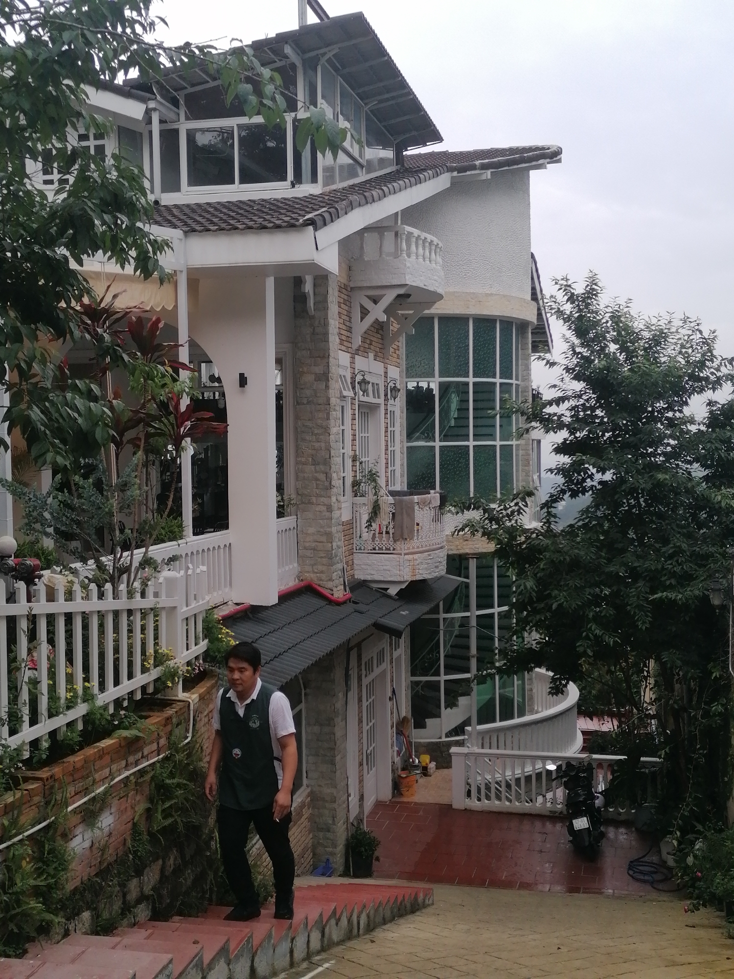 The image depicts a multi-story building, likely a house or small hotel, nestled on a slope amidst lush greenery in Da Lat, Vietnam, under an overcast sky. The building exhibits a distinctive architectural style with white plastered walls, some sections clad in light-colored stone, and dark tiled roofs. It features numerous windows, including large, grid-patterned glass panels enclosing an internal spiral staircase on the right side of the structure. Several white balconies with intricate railings extend from different levels.
In the foreground, a man walks up a set of red-tiled outdoor stairs. He wears a white short-sleeved polo shirt, a dark green vest with a patch, and black trousers. To his right, a white picket fence borders a garden area with a brick retaining wall covered in ferns and other plants, including some yellow flowers.
Various trees and potted plants surround the building, contributing to a verdant atmosphere. On the lower level, to the right of the man, a paved area with reddish-brown tiles is visible, where a black motorcycle is parked. The motorcycle's license plate displays "66-L4 444.61". A white curved railing defines the edge of this paved space. Cleaning tools, such as a broom and bucket, are seen near a door on the building's ground floor. The scene suggests a tranquil, possibly resort-like setting.