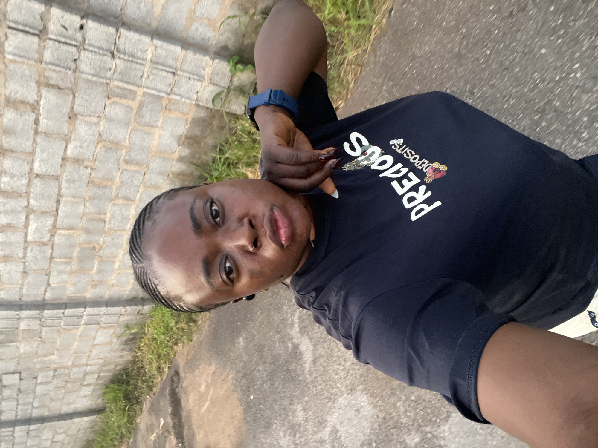 A young Black woman is taking a selfie outdoors. She has her hair braided and is wearing a dark blue t-shirt with white text and a small graphic on the front, and a blue smartwatch on her left wrist. She is resting her chin on her hand, and her lips are pursed. She is standing on a paved surface, with a grey brick wall behind her and grass growing along the base of the wall. The scene appears to be in an urban or semi-urban area, possibly in the city of Abuja, Nigeria, given the presence of paved roads, brick construction, and vegetation common in such environments.