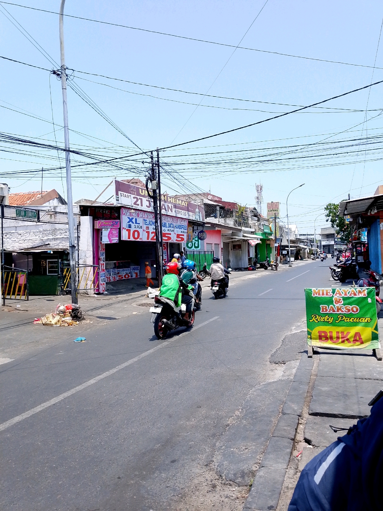 This image depicts a street scene in Surabaya, Indonesia, likely on a sunny day given the bright lighting and clear sky. The road is paved with asphalt and shows signs of wear, with cracks and uneven patches in places. A white line marks the center of the road. The scene is lined with various shops and buildings, typical of a commercial street.
On the left side of the road, a row of shops displays various signs and offerings. One prominent shop has a pink facade and several advertisements for mobile phone services and data packages, including "XL TELKOMSEL," "AXIS," and "3GB." Trash and debris are piled up near the curb in front of one of these establishments.
Several motorcycles are visible on the road. Two individuals are riding a motorcycle in the foreground, with the rider wearing a green vest, suggesting they might be a delivery person. Another motorcycle with two riders is seen further down the street. In the background, more motorcycles are parked or in motion, and a few pedestrians are present.
A bright green and yellow banner on the right side of the road advertises "MIE AYAM & BAKSO Rizky Pacuan BUKA," indicating a food establishment is open for business. The sidewalk is made of concrete slabs, some of which are cracked or uneven. Overhead, a complex network of electrical wires and poles crisscrosses the sky, a common sight in many urban areas. The overall impression is that of a busy, everyday street in an Indonesian city.