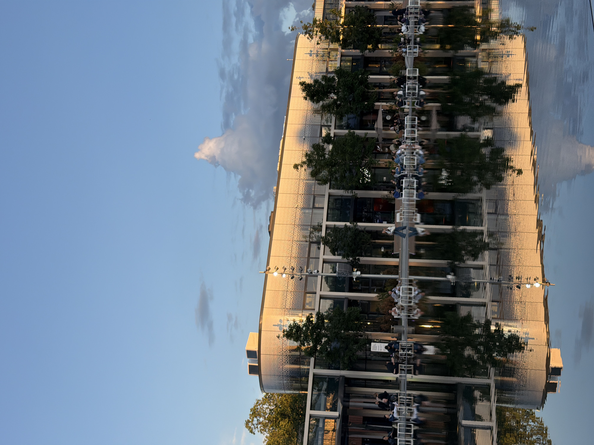 The image depicts a contemporary building with a curved facade, featuring a textured pattern that reflects the warm golden light of what appears to be a sunset. The building is set against a clear blue sky with some scattered clouds. In front of the building, there is a large body of water that acts as a reflective surface, mirroring the architecture, trees, and sky. Several people are seated at tables outside the building, seemingly enjoying an outdoor dining or cafe experience. The scene is tranquil, suggesting a public space or a commercial area designed for leisure. The reflection in the water adds depth and symmetry to the composition.
