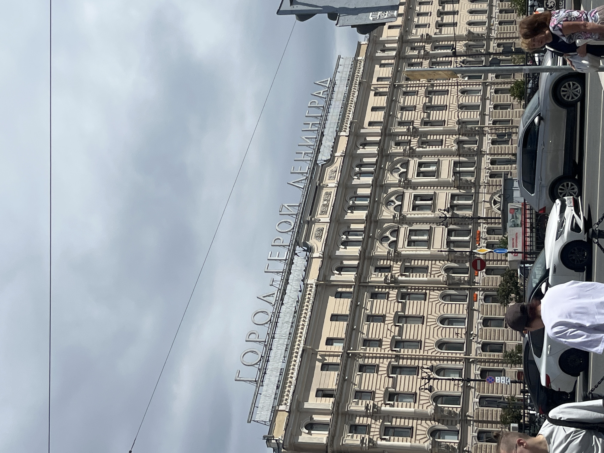 This is a photograph of the "Hero City Leningrad" sign on a building in Saint Petersburg, Russia. The sign is made of large, individual metal letters mounted on the rooftop of a multi-story building. The building is light-colored, with a classical architectural style featuring columns and arched windows. In the foreground, there is a street with cars and pedestrians. The sky is overcast with clouds. The image is taken from street level looking up at the building.