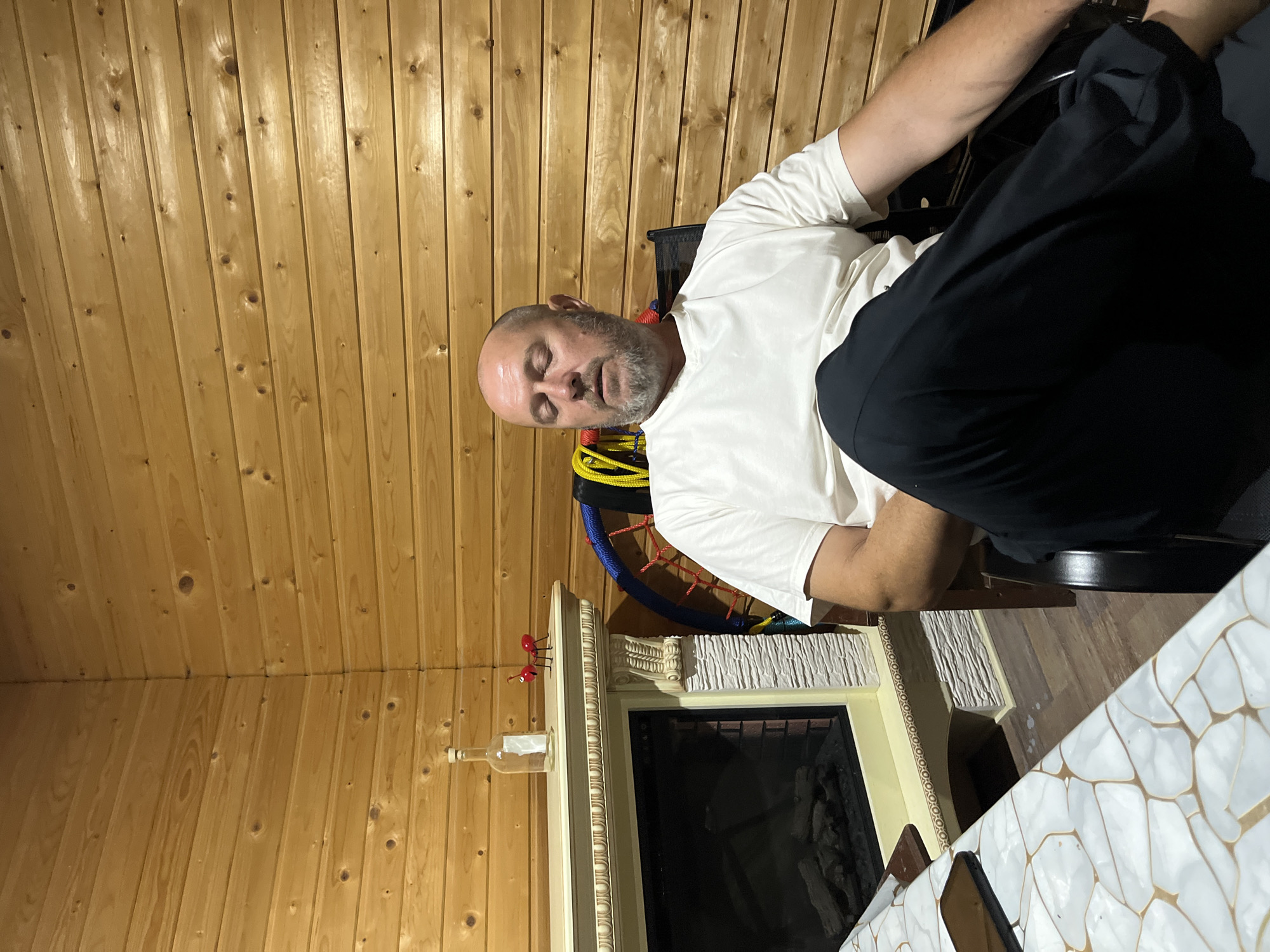 A bald, middle-aged Caucasian man with a grey beard is sitting in a chair with his eyes closed, appearing to be asleep or resting. He is wearing a white t-shirt and black pants. The man is in a room with wood-paneled walls. In the background, there is a fireplace with a decorative mantelpiece. On the mantelpiece, there is a clear glass bottle and two red decorative ant figures. To the right of the fireplace, a portion of a colorful woven structure is visible. In the foreground, a table with a white, patterned surface is partially visible. The lighting suggests an indoor setting, possibly a home or lodge. The image does not provide any information that would allow for its location in Saratov, Russia, to be determined.