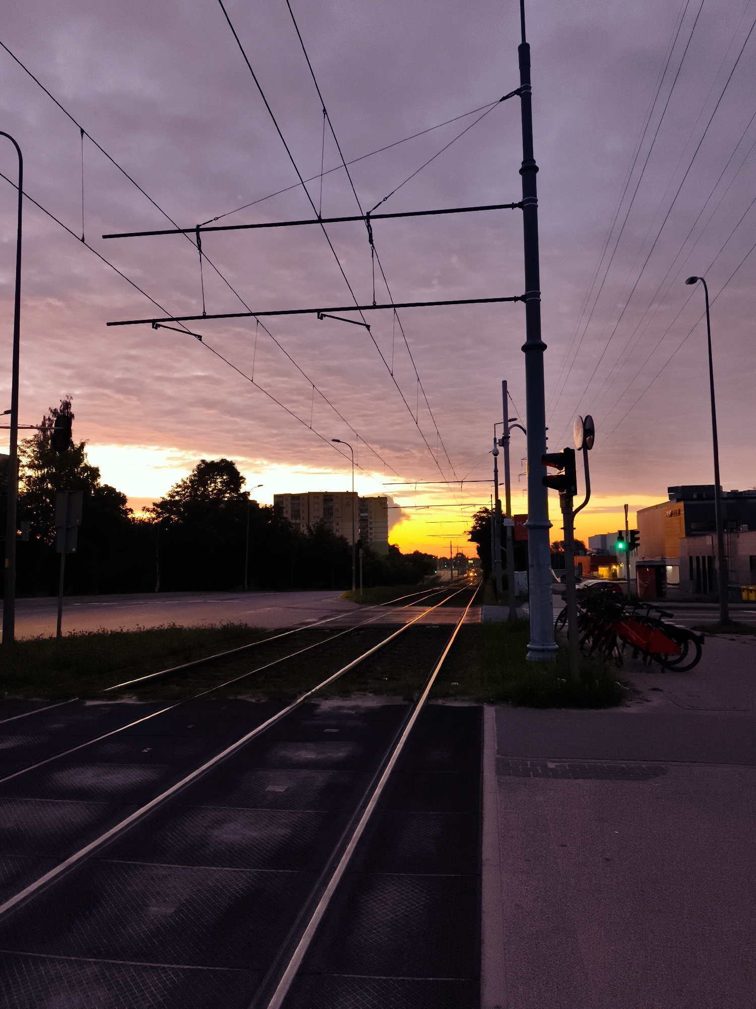 The image depicts a street scene in Gdańsk, Poland, at dusk. Tram tracks run diagonally across the foreground, disappearing into the distance. Overhead wires for the tram are visible against a purplish-pink sky filled with scattered clouds. A traffic light displaying a green signal is mounted on a pole to the right, along with a red traffic light for trams, indicating an intersection. Parked bicycles are gathered near the pole. In the background, silhouetted trees and buildings line the street. The setting sun casts a warm orange glow on the horizon. The overall impression is one of a quiet urban evening.