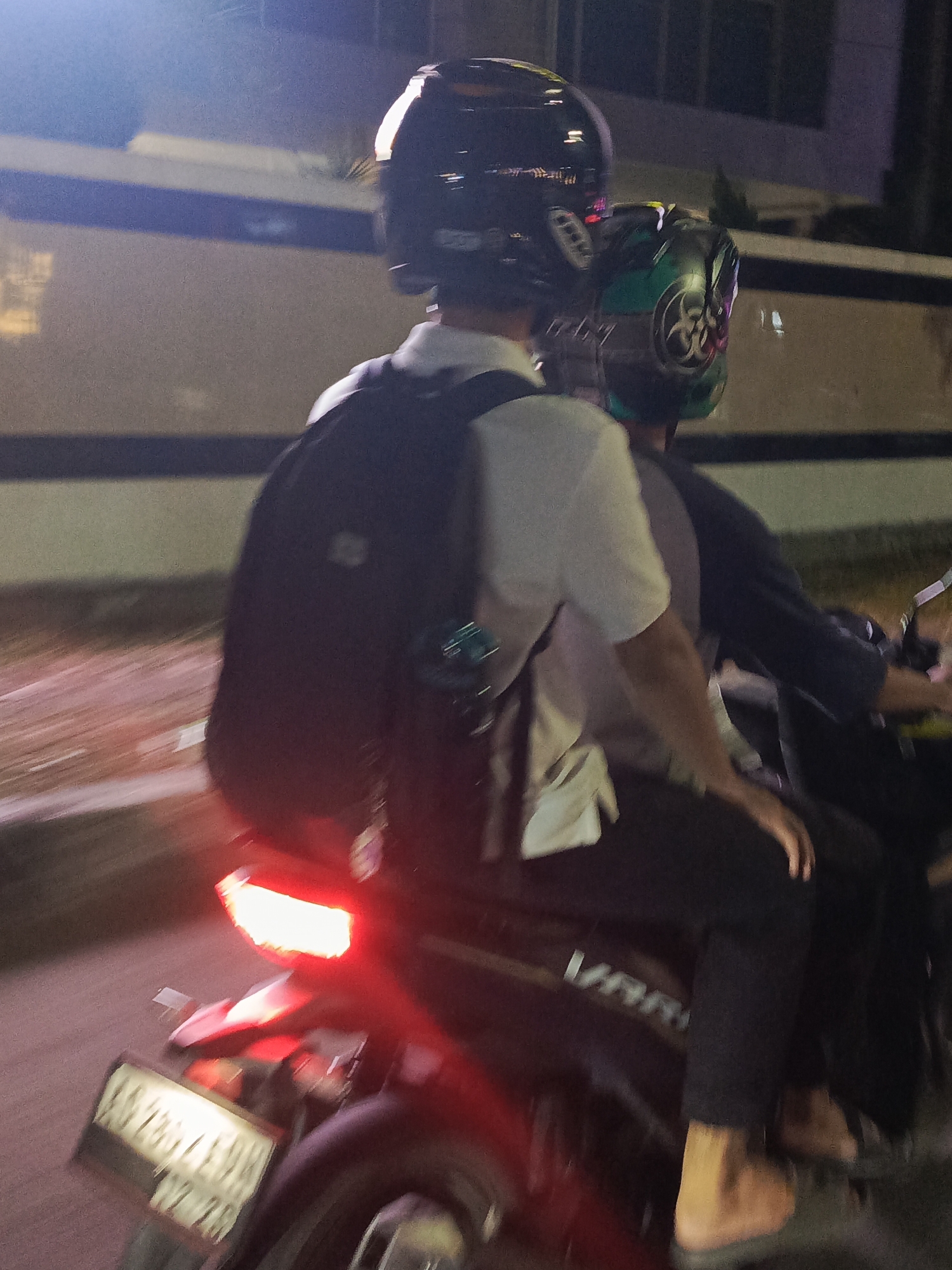 Two individuals are riding a motorcycle at night in Surabaya, Indonesia. The person in front is wearing a black helmet and a white short-sleeved shirt with a black backpack. The person in the back is wearing a green and black helmet. The motorcycle's taillight is illuminated red, and a license plate is visible at the rear. The background suggests an urban environment with blurred lights and structures. The motion blur indicates the motorcycle is in motion.