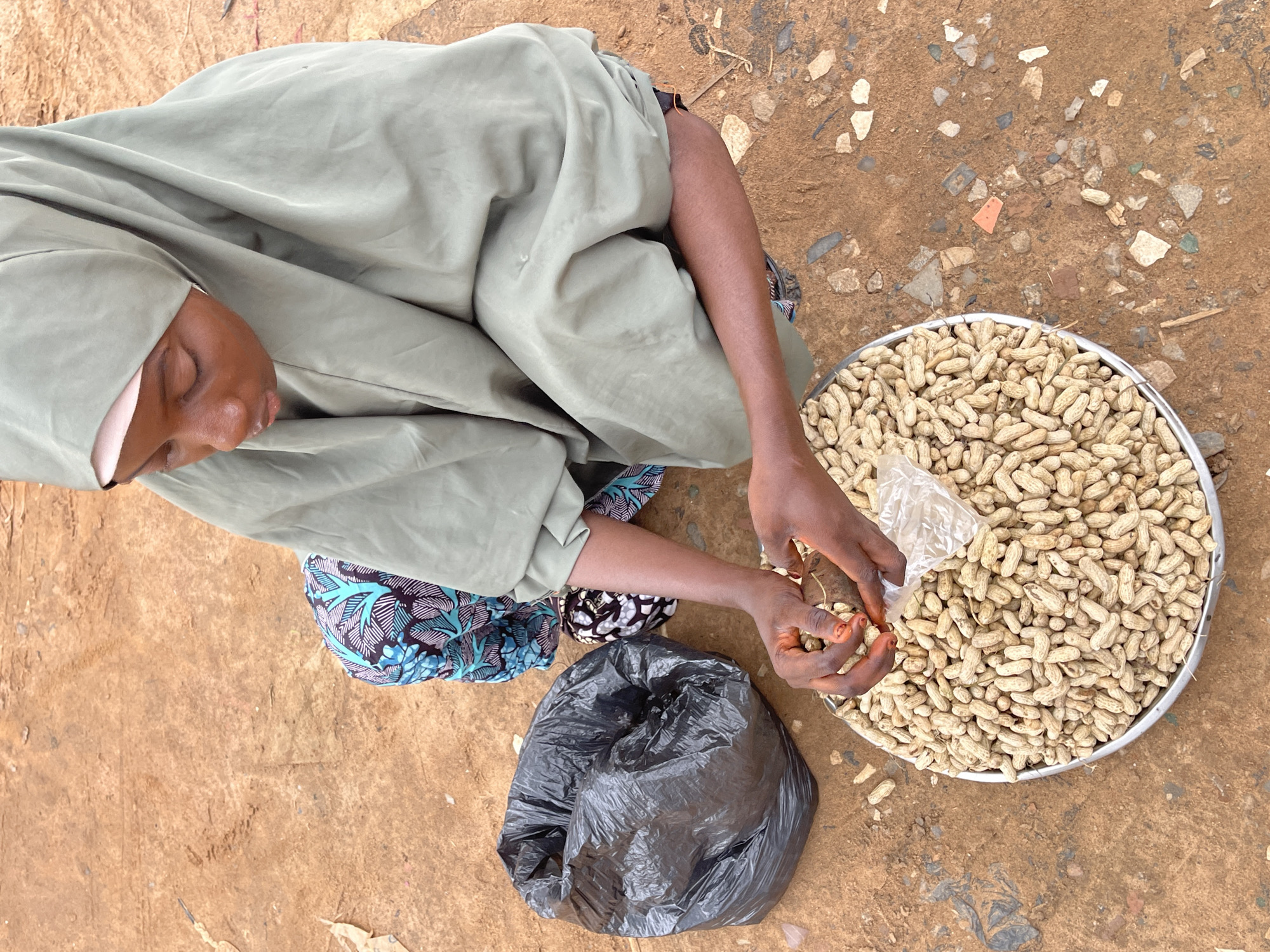 In Abuja, Nigeria, a person wearing a light green hijab and a colorful patterned skirt is crouched down on a dirt ground. In front of them is a large, round metal tray overflowing with peanuts in their shells. The person's hands are actively engaged in handling some of the peanuts, with a small clear plastic bag also present. To the left of the tray, a crumpled black plastic bag sits on the dirt. The environment appears to be an outdoor setting, possibly a market or street, characterized by the earthy brown soil with scattered small stones and debris.