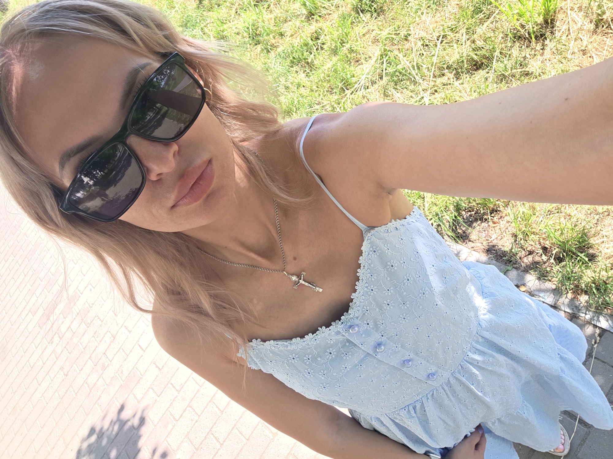 A woman is taking a selfie outdoors in Chaikovsky, Russia. She is wearing a light blue dress with white embroidery, a silver necklace with a cross pendant, and dark sunglasses. Her hair is blonde and wavy. She is standing on a paved area with visible brickwork, next to a grassy area. The lighting suggests it is daytime.