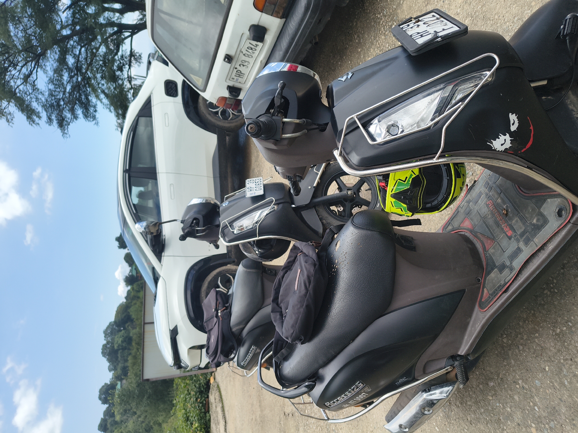 The image shows a parking area with several scooters and two cars. In the foreground, a black scooter is parked, with a black backpack placed on its seat. Another black scooter is parked behind it, also with a backpack on the seat. Further back, a white car and a white vehicle are visible. The environment appears to be outdoors, with a cloudy sky and trees in the background. The license plates visible on the vehicles suggest the location is in India. Specifically, one license plate reads "HP 39 G0653" and another reads "HP 39 6484". These license plates are indicative of Himachal Pradesh, a state in India. The overall scene depicts a typical day in a parking lot, likely in a rural or semi-urban area within India.