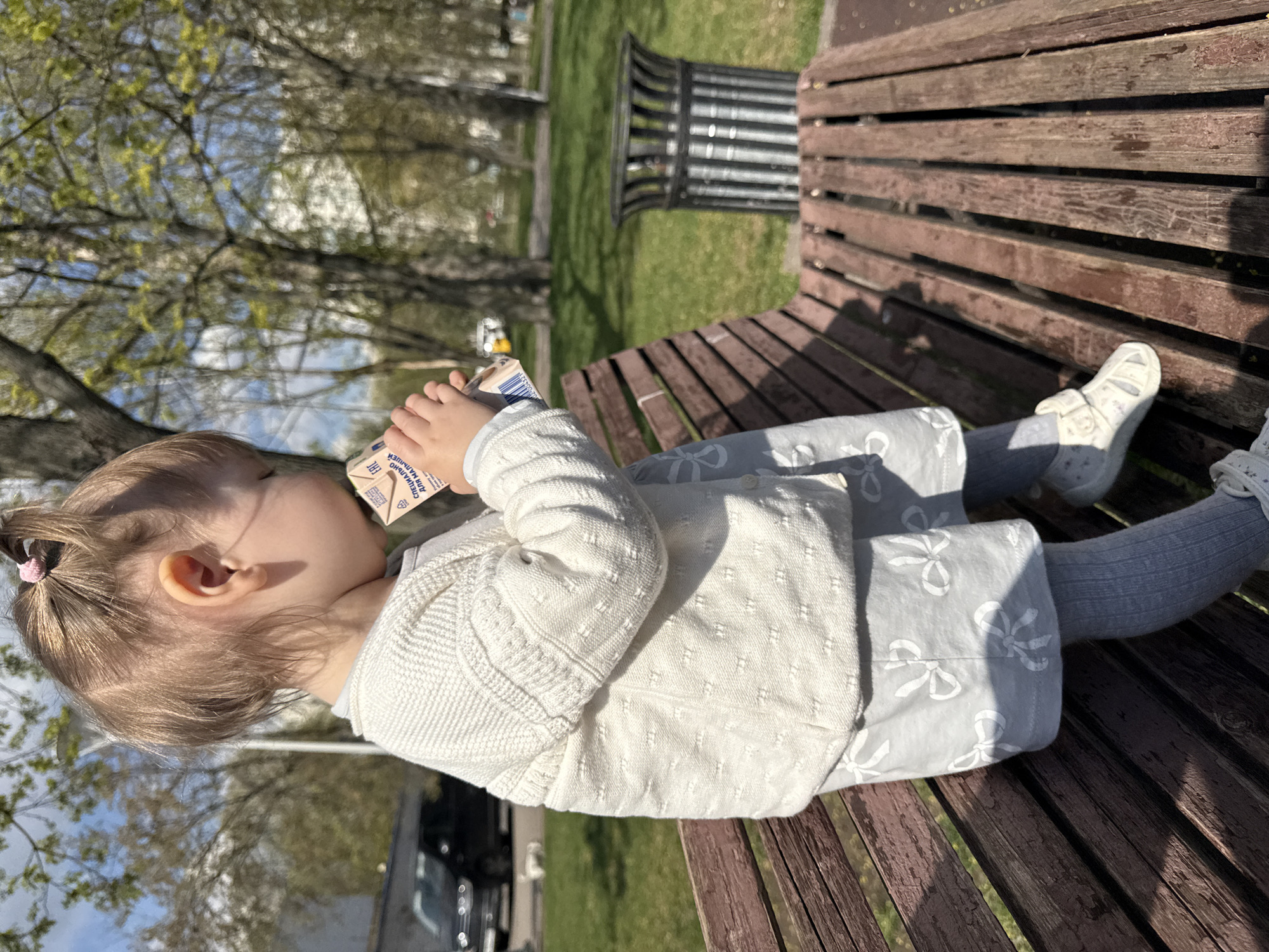 A young child is sitting on a wooden park bench, holding a small carton of a beverage, possibly juice or milk, to their mouth. The child is wearing a light-colored knit sweater, grey patterned leggings or pants, and light-colored shoes with velcro straps. The park bench appears to be in a public park with greenery and trees in the background, along with a metal trash receptacle near the bench. The photograph was taken outdoors during daylight hours. The beverage carton contains Russian text. It is likely located in Moscow, Russia.