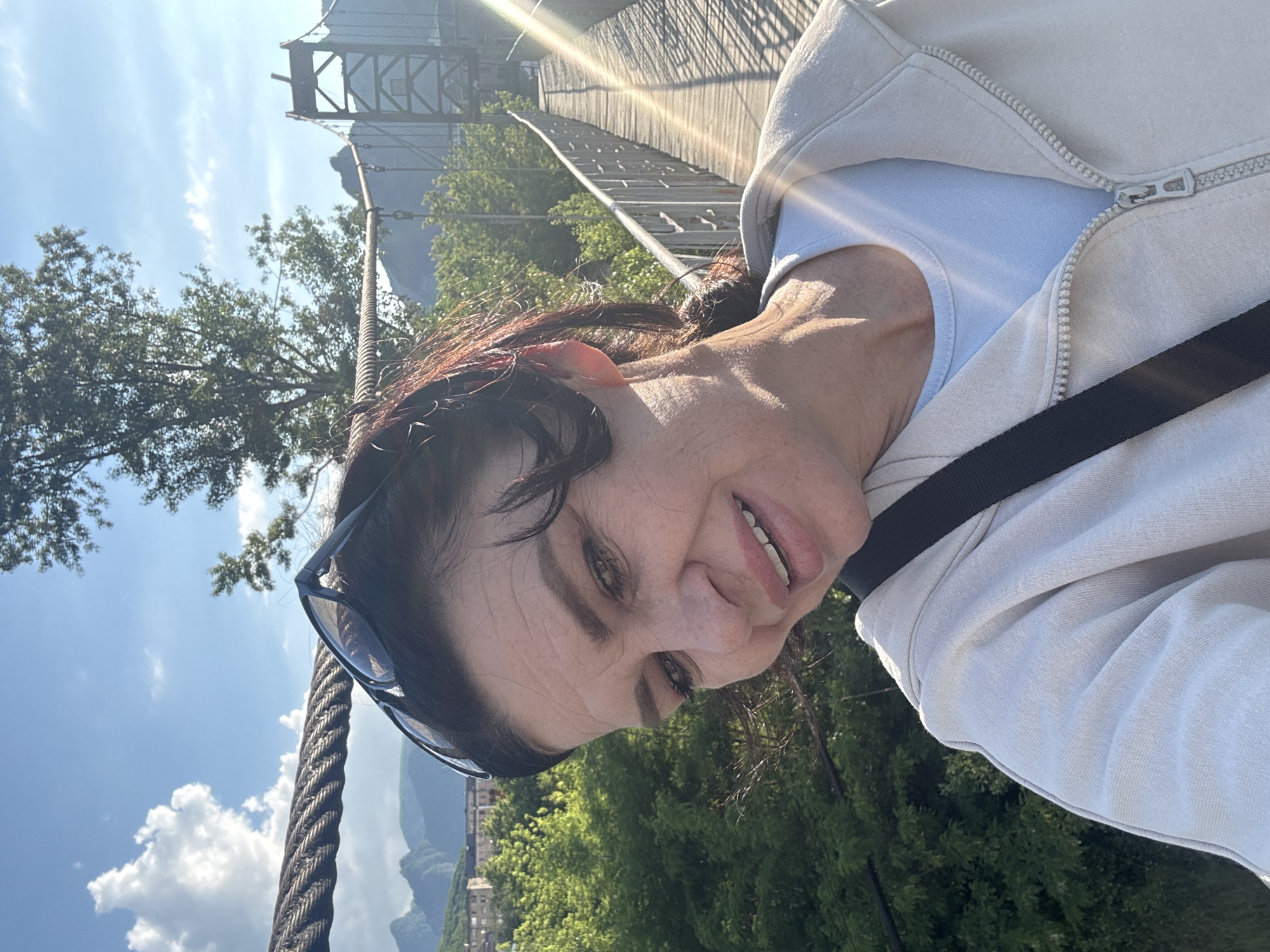 A woman with dark hair and sunglasses on her head takes a selfie on a suspension bridge. She is wearing a light-colored zip-up hoodie over a light blue t-shirt. The bridge has wooden planks and metal railings. In the background, there are green trees, mountains, and buildings. The sun is shining brightly, casting lens flares into the image. This bridge is likely the suspension bridge over the Kuban River in Karachayevsk, Russia, which is known for its scenic views of the surrounding Caucasus Mountains.