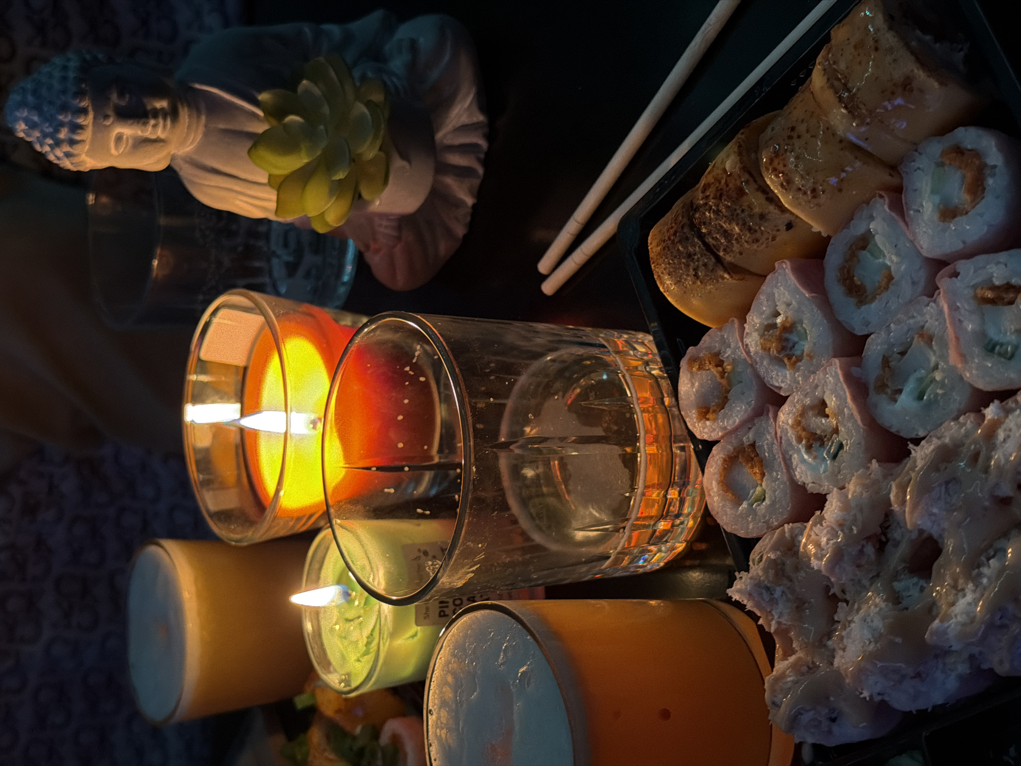 The image depicts a serene, low-light indoor setting, likely an evening scene in Varna, Bulgaria, illuminated predominantly by candlelight. The primary subjects include a variety of sushi or maki rolls, candles, drinks, and a decorative Buddha figurine.
On the right side, a black tray holds an assortment of sushi. Some rolls have a golden-brown, possibly seared or baked topping, while others feature a lighter, pinkish-white exterior with visible fillings. A significant portion of the sushi is topped with a creamy white sauce or finely shredded ingredients. A pair of light-colored chopsticks rests on the tray, indicating an ongoing meal.
The scene is lit by several candles. Two are prominent in the foreground, housed in clear glass holders; one emits a warm orange glow, and another a softer, greenish-yellow light. Two larger cylindrical candles are also present, one in a light orange-brown hue and another with a light blue top surface. A clear glass tumbler, partially filled with a transparent liquid, is situated near the candles.
In the upper left, a white, possibly ceramic or stone, Buddha figurine is visible, adorned with a small green succulent plant on its shoulder. The overall atmosphere is intimate and tranquil, suggesting a relaxed dinner or a moment of quiet enjoyment. The lighting emphasizes warm tones and creates a cozy ambiance against a dark background. Faint, partially obscured text, possibly "PILL", can be seen on the side of one of the candle holders.