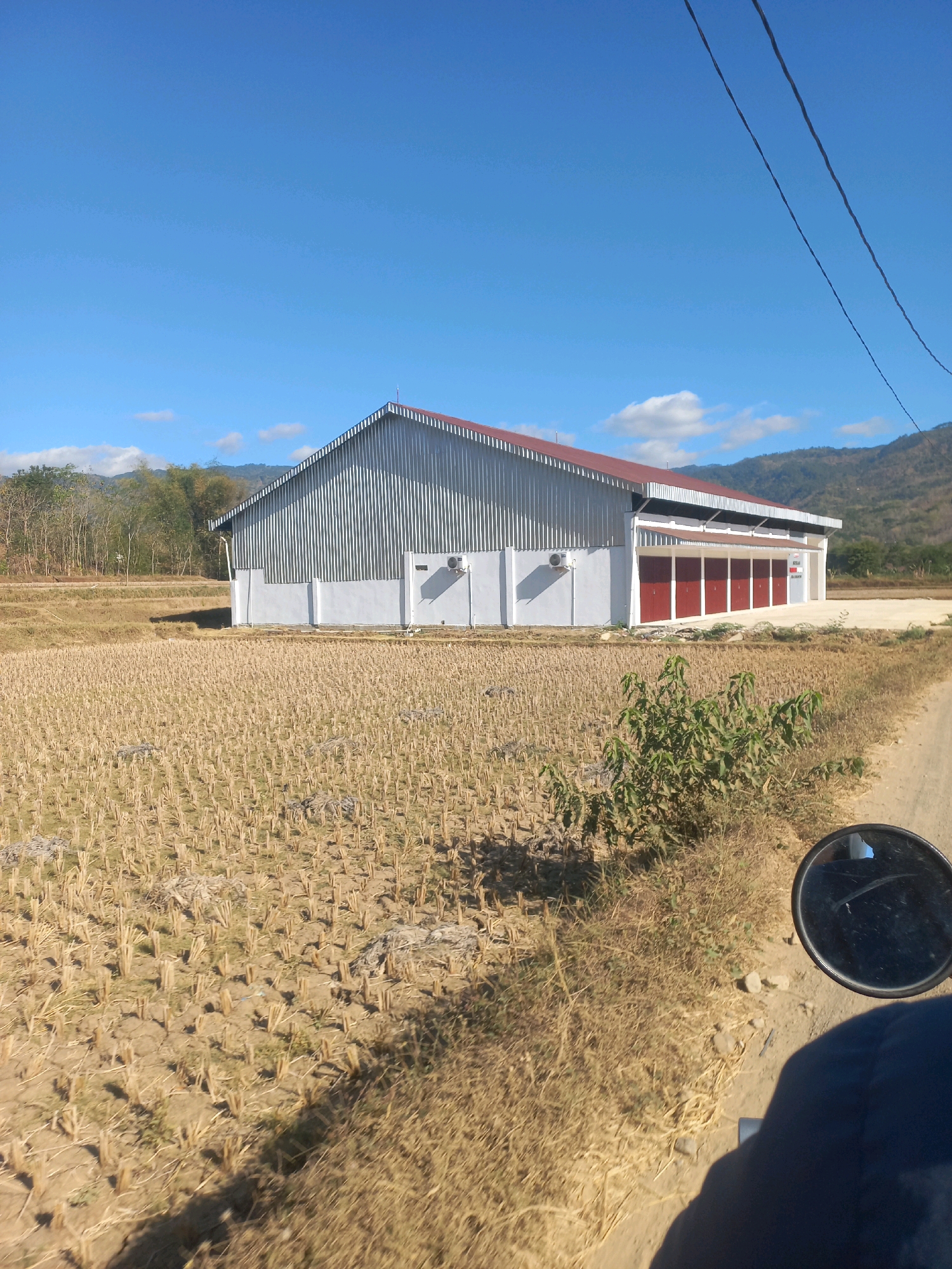 The image depicts a rural scene in Girirejo, Indonesia, featuring a modern, corrugated metal building with a red roof, situated in front of a harvested rice field. The field is dry and stubbled, with rows of remaining rice stalks visible. The building has a white base and several red doors. Two air conditioning units are mounted on the white wall of the building. In the foreground, a dirt road is visible, along with the rearview mirror and a portion of a dark blue garment, suggesting the photograph was taken from a vehicle. Overhead power lines stretch across the bright blue sky, which is dotted with a few white clouds. In the background, a line of trees and hills can be seen. The overall impression is one of agricultural activity and modern infrastructure in a natural, hilly landscape.
