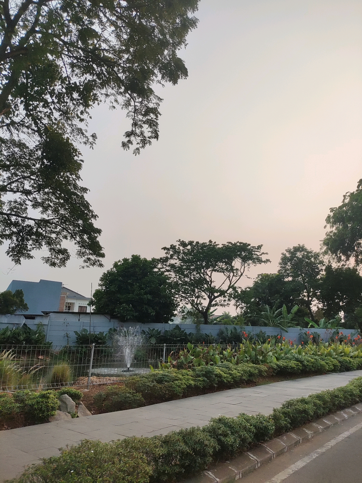 The image shows an outdoor scene in Telagaasih, Indonesia, featuring a paved walkway bordered by lush green bushes. Beyond the walkway, a fence encloses a garden area with a fountain spraying water. Several trees are visible, with dense foliage casting shadows. In the background, a light blue building and a wall can be seen. The sky is overcast with a pale, hazy light. The overall impression is a well-maintained public space, possibly a park or a residential area's common grounds.