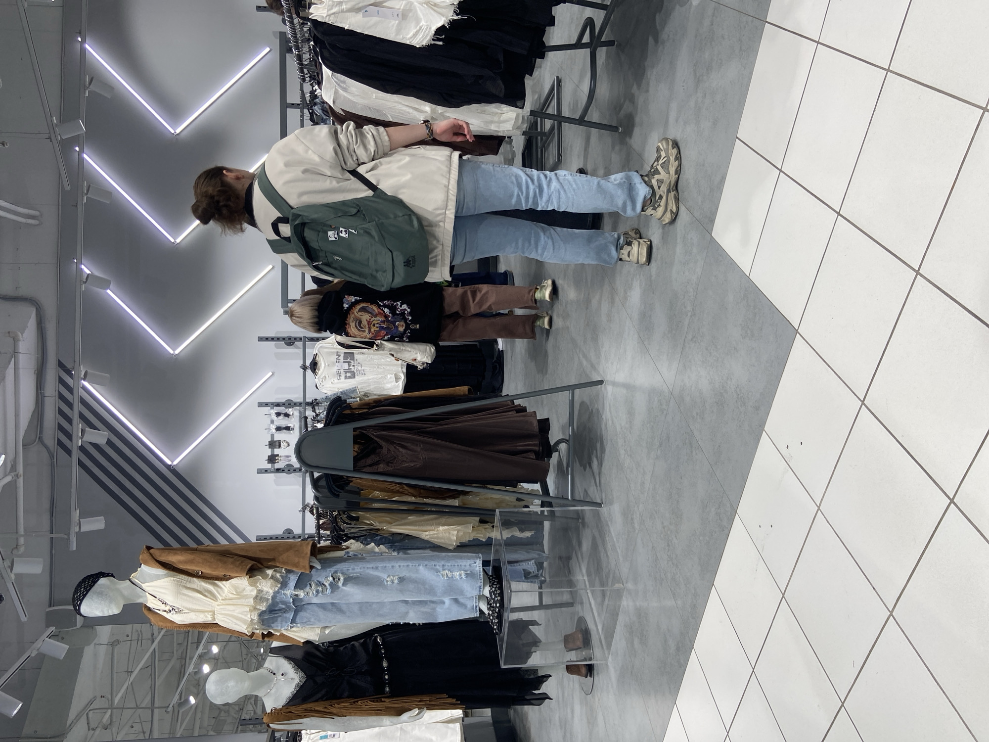 The image depicts an indoor scene within a modern clothing store, likely in Saint Petersburg, Russia. Two individuals are central to the frame, viewed from behind. The taller person, an adult, has light brown hair tied in a bun and is dressed in a light-colored bomber jacket, light blue jeans, and chunky, multi-toned sneakers. They wear a dark green backpack adorned with a small white rectangular badge and a round dark pin. Standing next to them is a smaller individual, possibly a child, wearing a black long-sleeved shirt with a prominent colorful graphic design on the back, brown pants, and light-colored sneakers. Both individuals appear to be browsing clothing displayed on metal racks.
The store's interior features several clothing racks stocked with various garments, including white shirts, black dresses, brown jackets, and denim jeans. Mannequins, dressed in contemporary casual attire such as ripped jeans, a cream-colored top, and a brown suede-like jacket, are visible in the foreground. The floor is covered with large tiles, transitioning diagonally from a dark grey on the left to white on the right. The ceiling is equipped with linear fluorescent lights arranged in V-shapes, providing bright illumination, alongside exposed pipes and air ducts. A clear acrylic display stand is positioned on the floor near the mannequins. No explicit brand names or readable text are clearly visible on any clothing items or objects, though the child's shirt has an intricate graphic and a mannequin's shirt has partially obscured text. The overall ambiance suggests a retail environment.