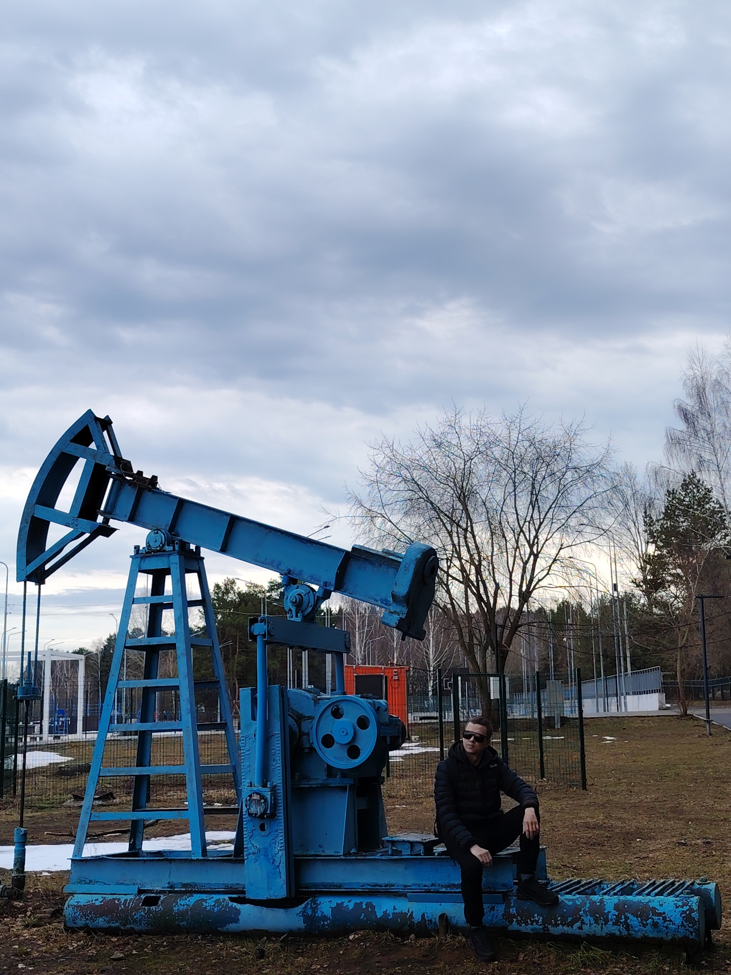 A medium shot shows a man sitting on a blue-painted oil well pumpjack in Chaikovsky, Russia. The man, wearing dark sunglasses and a black puffer jacket, is positioned to the right of the pumpjack. The pumpjack is weathered with chipped paint, and is surrounded by a sparse, grassy area with patches of melting snow. Bare trees and a building with a wire fence are visible in the background. The sky is overcast with gray clouds.