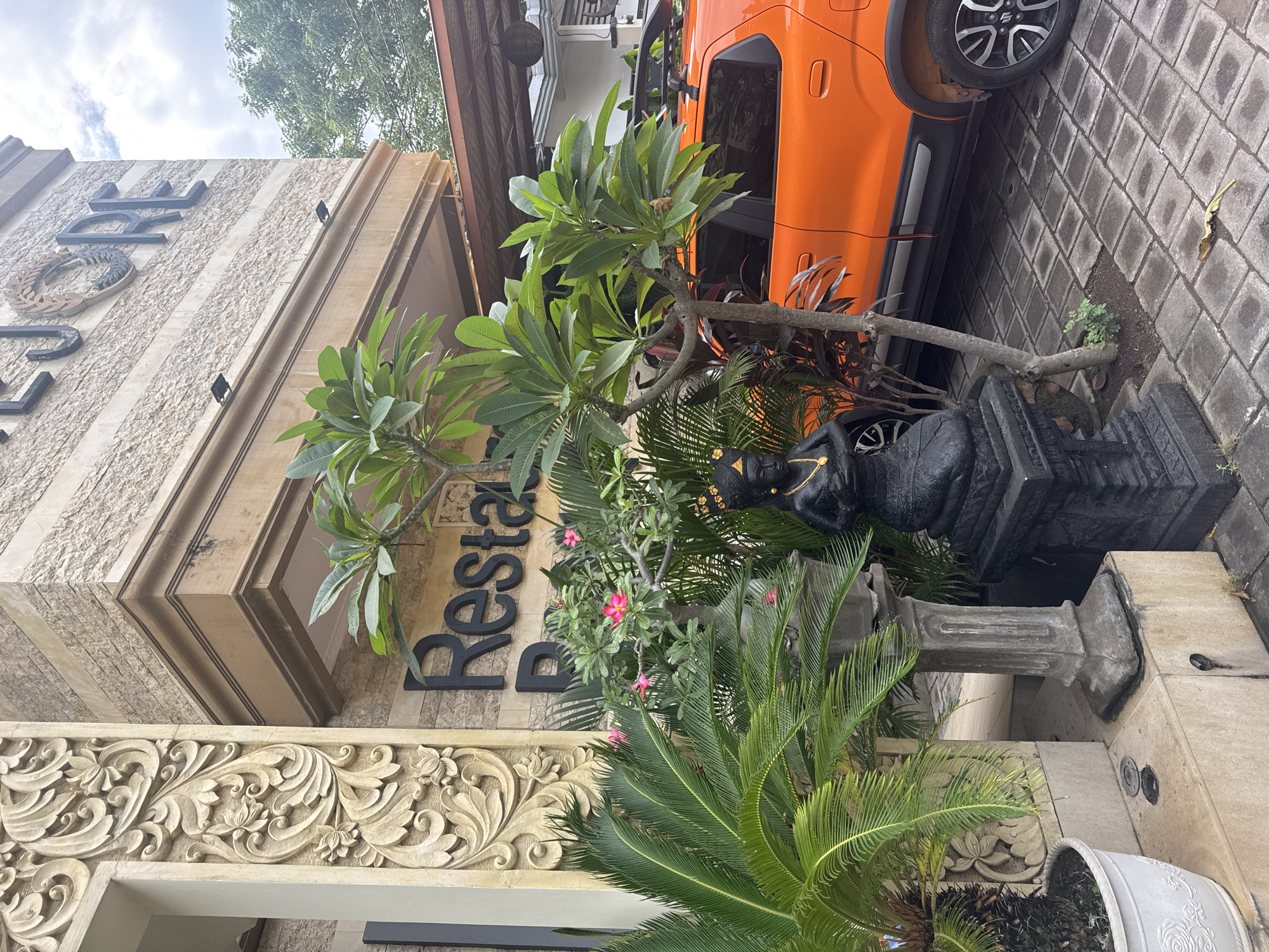 The image captures an outdoor scene in front of a building with the name "JORE" visible in large black letters on the facade. The building features decorative stone carvings on its left side and a patterned stone exterior. Below the name, the word "Restau" is partially visible.
In the foreground, there are several lush green plants, including a large palm with fronds extending to the left and a smaller tree with broad leaves and pink flowers. A black statue of a kneeling figure with a headdress and necklace is positioned among the plants. To the right of the plants, the rear of an orange car is visible, parked on a paved surface made of dark gray square tiles. The car has a roof rack and its rear tire is visible. The environment appears to be a public space, possibly an entrance to a restaurant or establishment. The lighting suggests an overcast day.