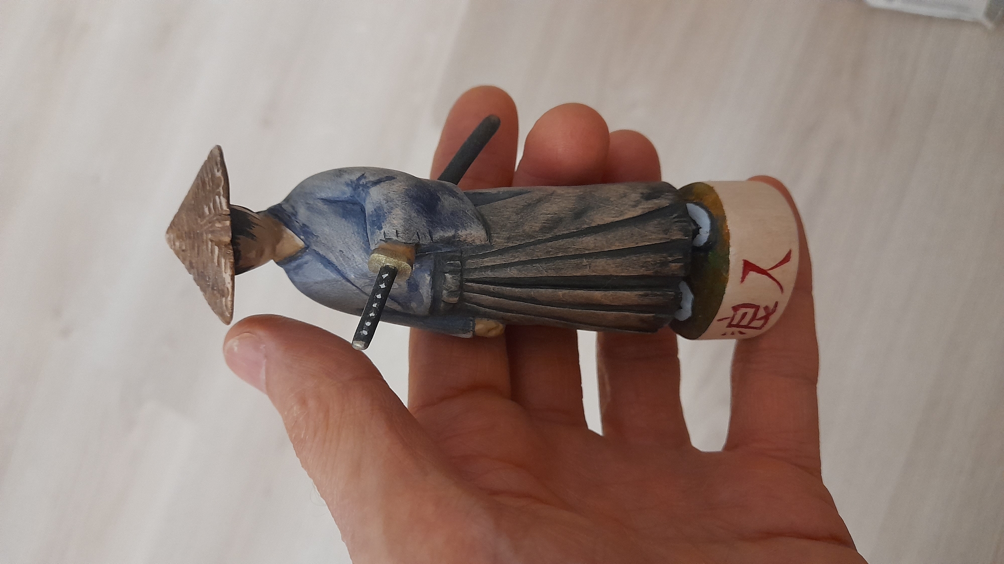 A hand holds a small, hand-painted wooden samurai figurine. The figurine is dressed in a blue kimono and a conical hat. A sword is visible at its side. Japanese characters are painted on the base of the figurine. The background is a light-colored wooden floor. The location appears to be indoors. There is no evidence this image was taken in Saint Petersburg, Russia.