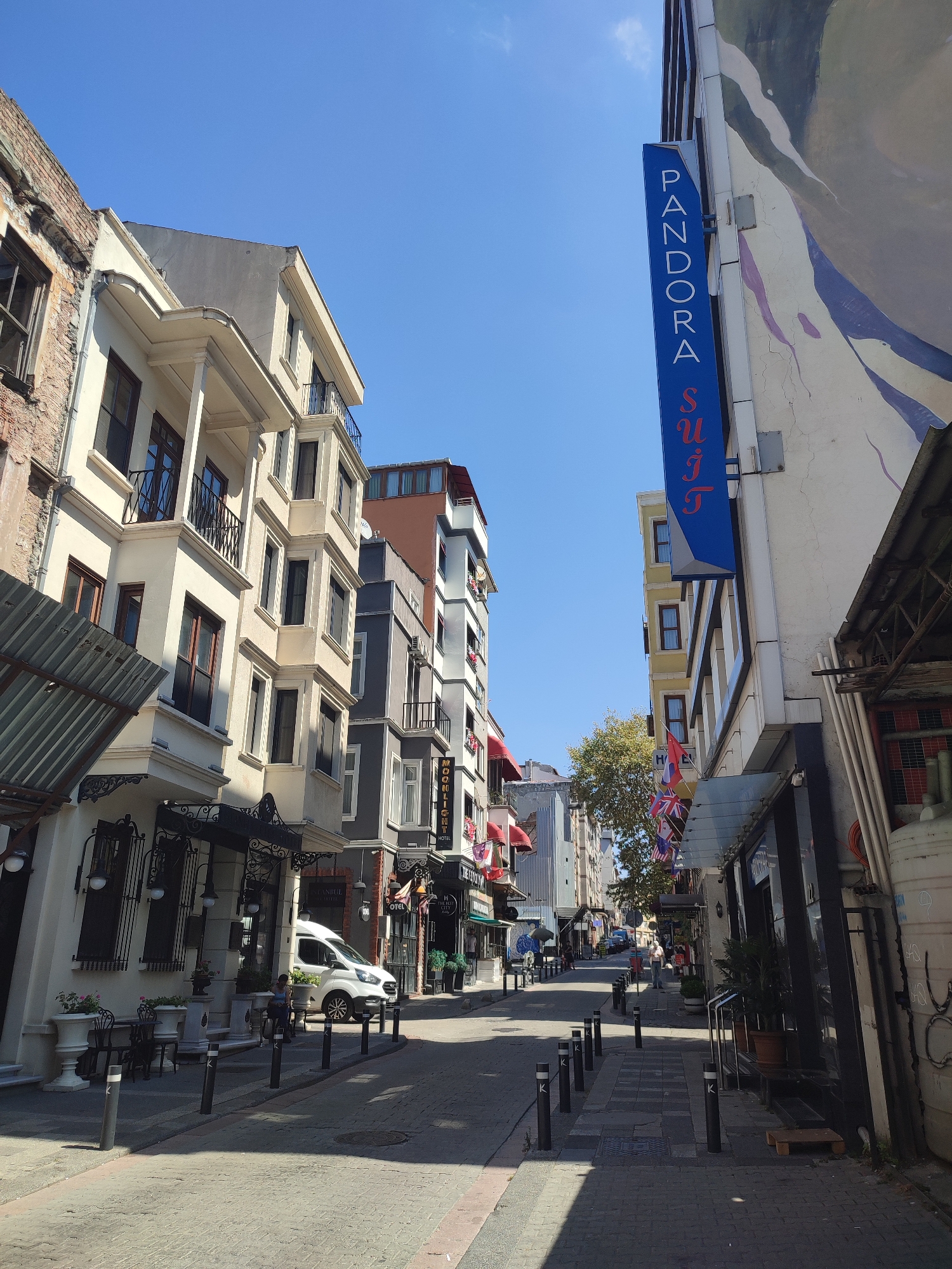 This image captures a street scene in Kadıköy, Turkey, on a sunny day. The perspective is from the middle of the street, looking down a narrow lane lined with multi-story buildings.
On the left, a light-colored building with ornate balconies and dark-framed windows dominates the foreground. Adjacent to it, a partially visible brick building shows signs of age. The building on the right is taller and modern, featuring a large mural or painted advertisement on its facade and a prominent blue vertical sign that reads "PANDORA SUIT".
The street is paved with cobblestones and lined with black bollards. Several parked cars, including a white van, are visible further down the street. Pedestrians can be seen walking and standing on the sidewalks. The architecture suggests a mix of historical and contemporary styles, typical of urban areas. The sky is clear blue, indicating fair weather. Small potted plants and outdoor seating are present outside some of the establishments. Flags, including British Union Jacks, are displayed above a storefront on the right side of the street.