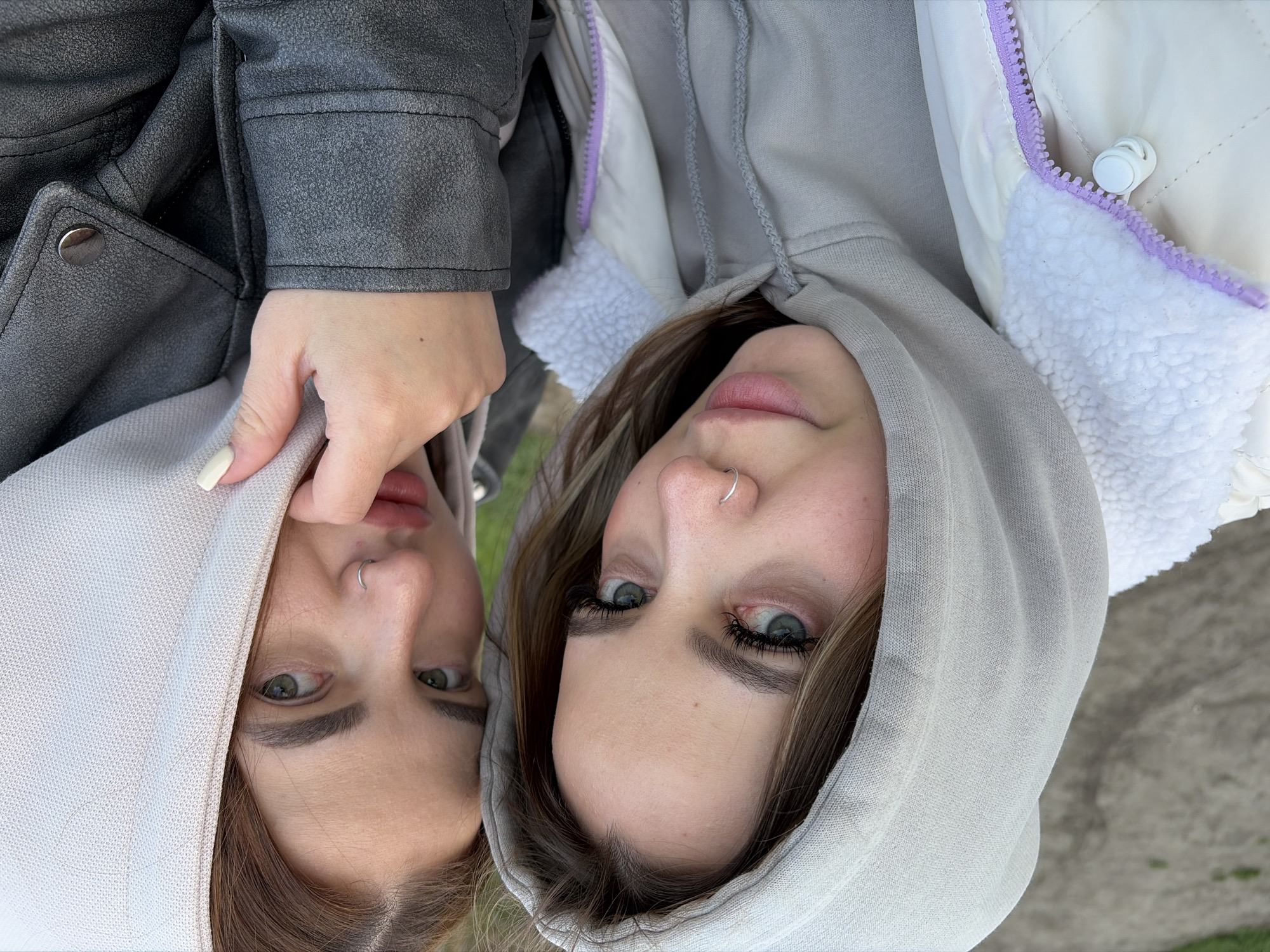 Two young women are pictured outdoors, their faces close together and angled towards the camera. The woman on the left wears a gray jacket and holds the hood of her light gray hooded sweatshirt with her left hand. The woman on the right wears a white jacket with a lavender zipper and white furry trim around the hood, which is pulled up over her head. Both women have nose rings. The location is indeterminate beyond being outdoors, there is no information to suggest they are in Krasnodar, Russia.