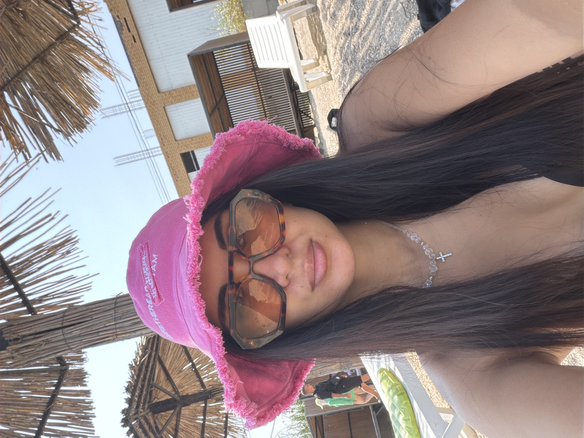 A medium shot captured from a low angle shows a woman wearing a pink bucket hat and large, tortoiseshell sunglasses. She is at the beach, as indicated by the sandy ground and a thatched umbrella overhead. Behind her, a partially constructed building with visible rebar stands against a clear sky. To the right, a white plastic lounge chair is positioned on the sand. In the background, two individuals are visible walking away from the camera, one in a black shirt and shorts, the other in a green top and shorts. A green inflatable object rests on a white lounge chair in the foreground. The woman is wearing a crystal necklace with a cross pendant.