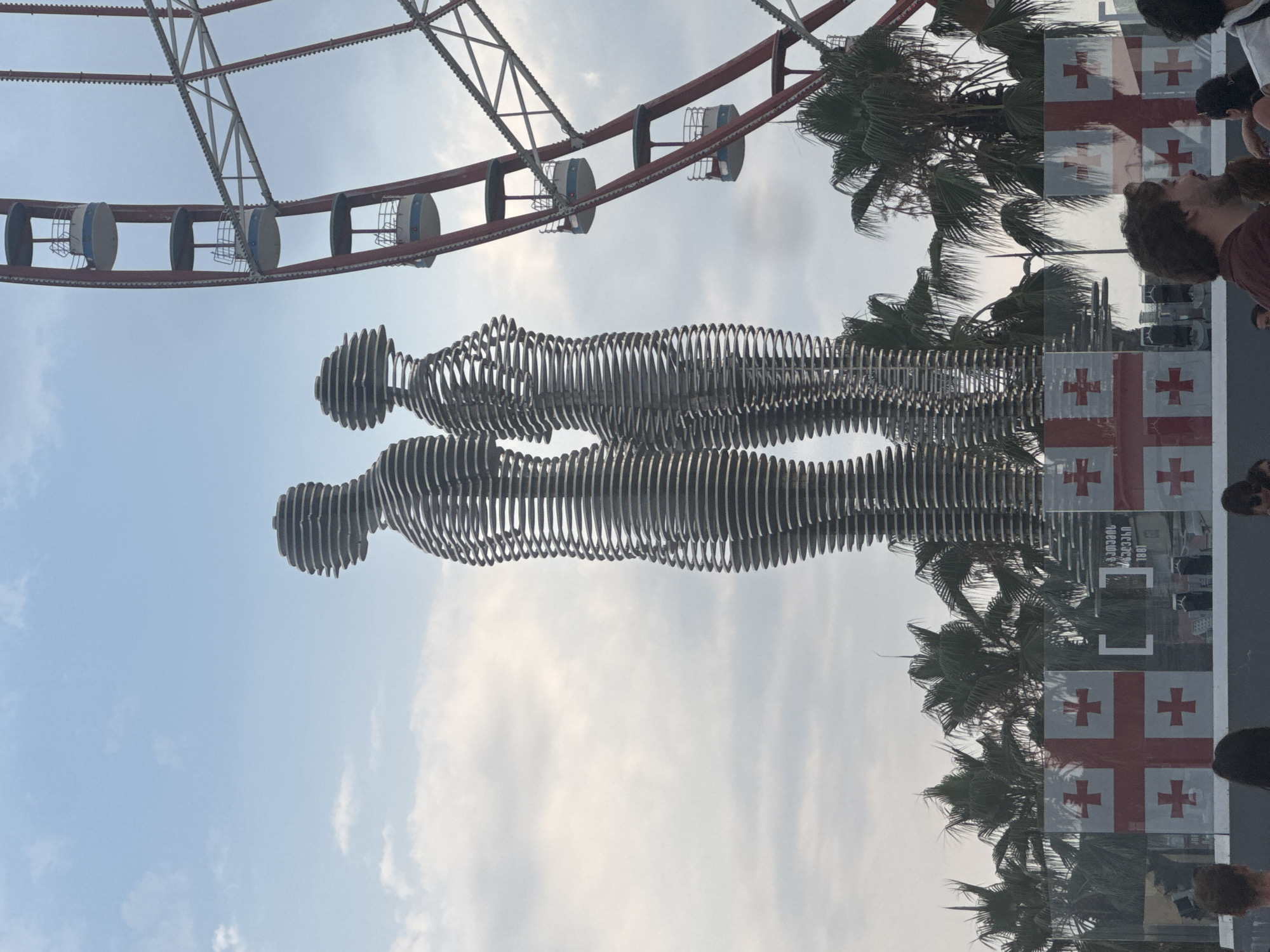 In Batumi, Georgia, a public monument featuring two interwoven metal figures, known as "Ali and Nino," stands against a backdrop of palm trees and a cloudy sky. A section of a Ferris wheel, with its red spokes and gondolas, is visible to the right of the sculptures. The base of the monument is adorned with glass panels displaying the Georgian flag. People are present in the foreground, some in profile, observing the scene. The overall setting suggests an outdoor urban environment.