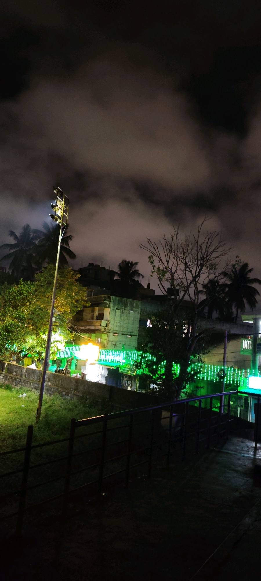 A nighttime outdoor scene in Belagavi, India, depicts a row of buildings illuminated by decorative green lights and a bright white light from a tall stadium-like spotlight. The sky is filled with dark, wispy clouds, suggesting a possibly humid or overcast night. Palm trees and other foliage are visible, some brightly lit by the artificial lights. In the foreground, a dark metal railing runs horizontally across the image, casting shadows on a paved surface. The overall impression is one of an urban nightscape with festive or functional illumination against a dramatic cloudy sky.