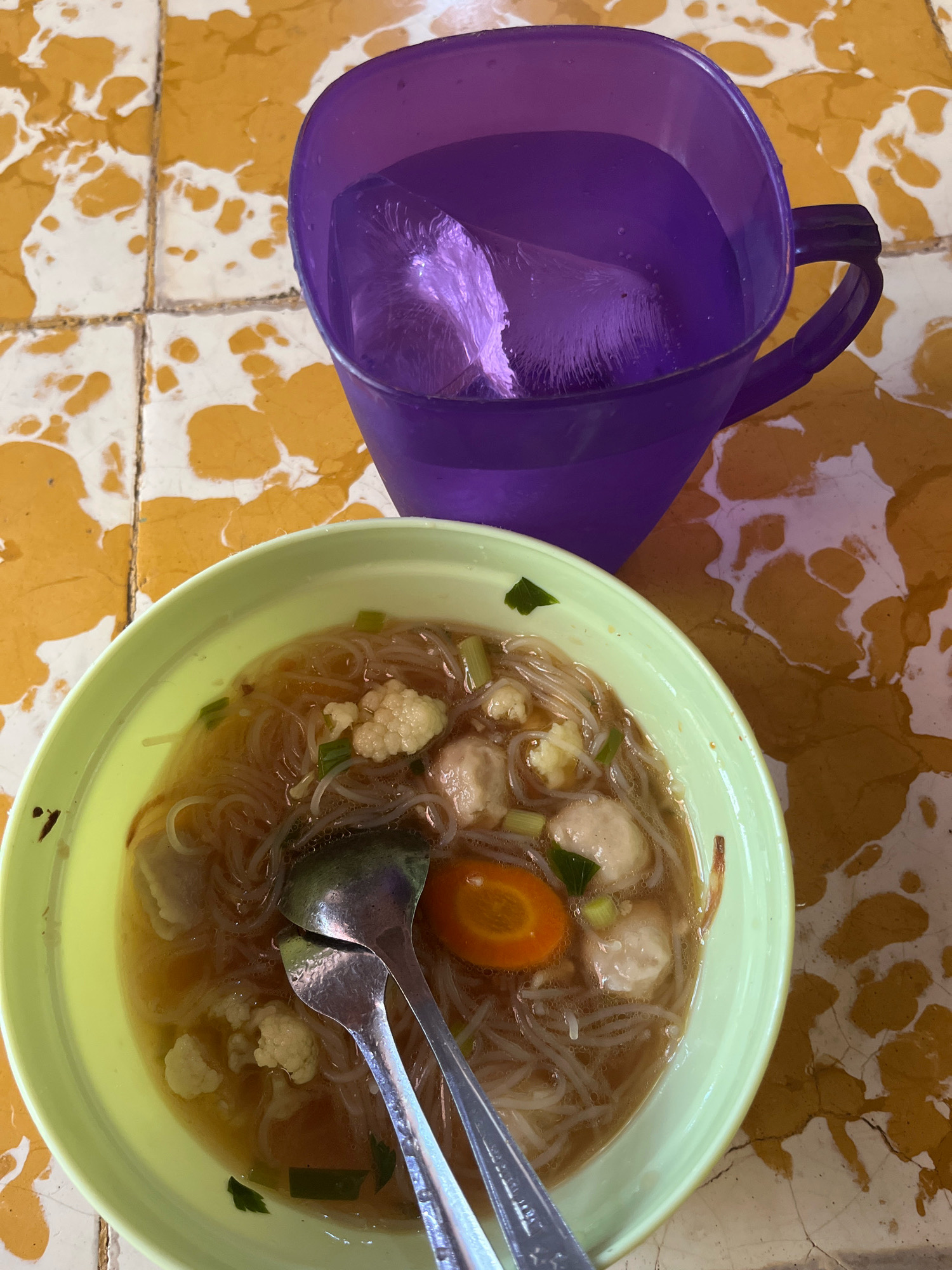 An overhead, close-up shot shows a bowl of soup and a cup of drink on a speckled yellow and white tiled surface, likely in Lambaro Kueh, Indonesia. The bowl, which is light green, contains a broth with noodles, cauliflower florets, meatballs, and a slice of carrot. Two silver spoons are resting in the soup. To the right of the bowl, a purple plastic cup holds a clear liquid with ice cubes. The tiled surface appears worn and possibly outdoors, given the dappled light and shadows.