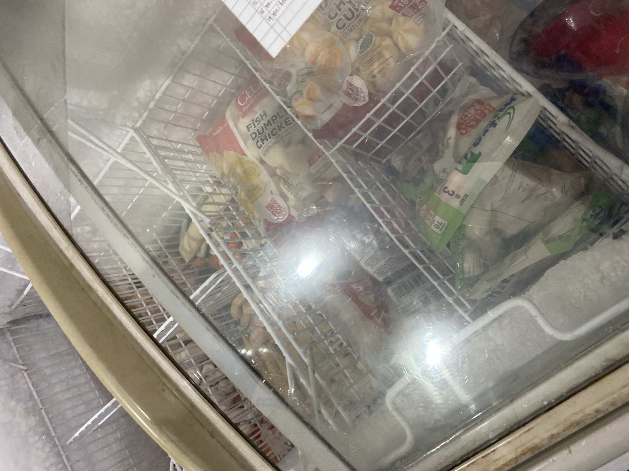 The image shows the inside of a commercial chest freezer, filled with various frozen food products organized in wire baskets. The freezer has a glass lid, which is fogged and has some condensation, obscuring a clear view of all the items. Visible products include bags of dumplings, such as "FISH DUMPLING CHICKEN" and "CHICKEN DUMPLING," and other food items labeled in Indonesian, like "BAKSO SAPI" which translates to beef meatballs. Some products have nutritional information and barcodes. A price tag or inventory sticker with handwritten text is attached to the edge of one basket. The overall environment appears to be a retail or food service setting, likely a store or restaurant in the city of Payatieng, Indonesia, due to the Indonesian labeling on the products. The freezer itself shows signs of use and cold storage, with visible frost on the interior walls. There are no people present in the image.