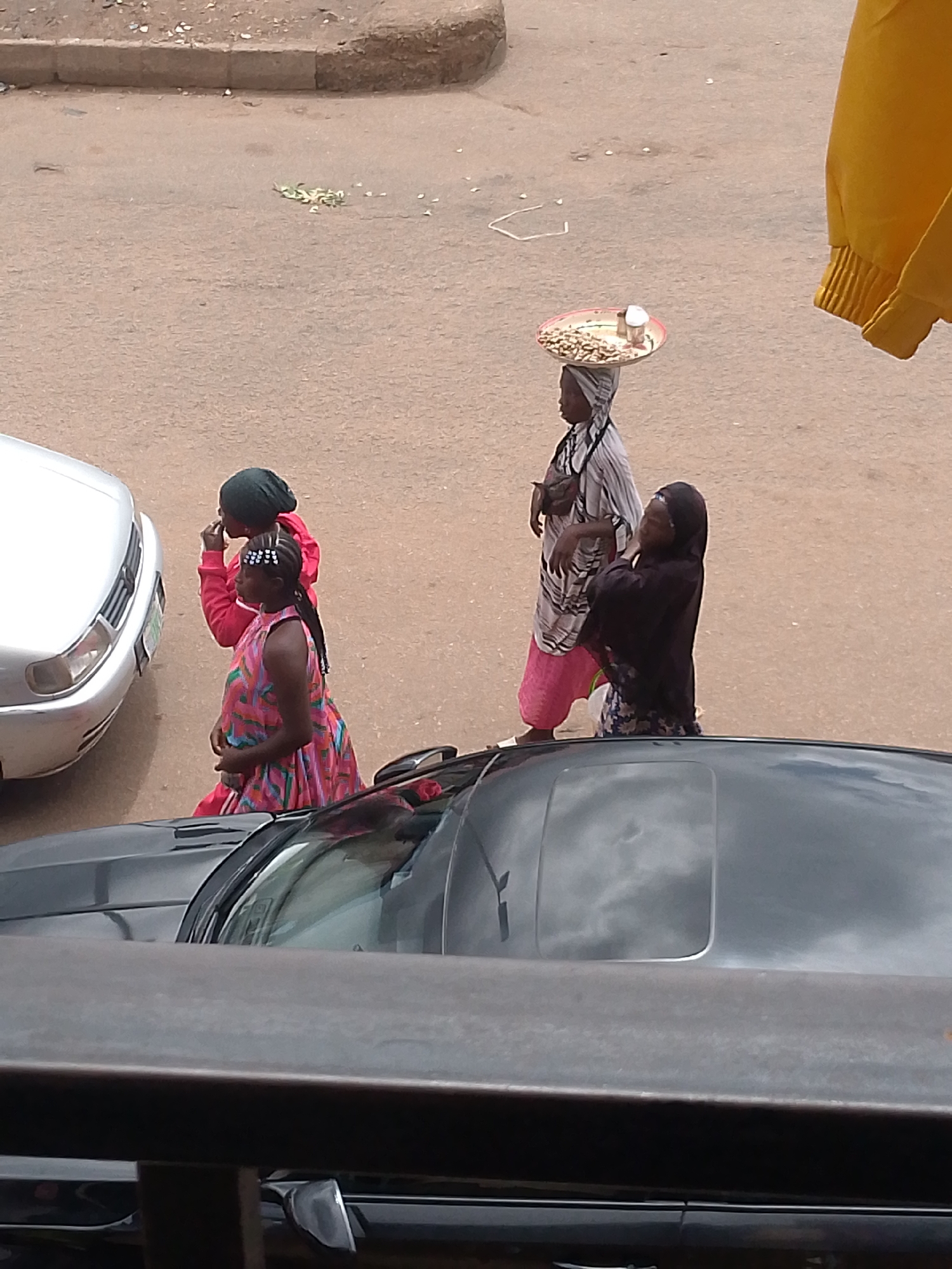 The image depicts an outdoor scene in Bukuru, Nigeria, featuring several individuals and vehicles on a dusty, paved surface that appears to be a street or market area. In the foreground, the top of a dark-colored car with a sunroof is visible, suggesting an elevated vantage point for the photograph. To the left, the front of a silver car is partially visible.
Three women are the central subjects. The leftmost woman is wearing a dark headscarf and a pink hooded jacket over a patterned dress. She is looking to her left and appears to be holding her hand to her mouth. To her right, a second woman, also wearing a headscarf, is walking and balancing a shallow tray on her head, laden with food items that resemble nuts and a small white object. She is wearing a striped top and a pink skirt. To the right of the second woman, a third woman is dressed in a dark headscarf and a dark garment, and she is looking upwards and to her right, with her hand near her neck.
Scattered on the ground, particularly in the upper left quadrant of the image, are small, light-colored items, possibly debris or discarded food. The ground surface is predominantly a reddish-brown dirt and pavement. A portion of a yellow garment is visible hanging from the top right edge of the frame.
