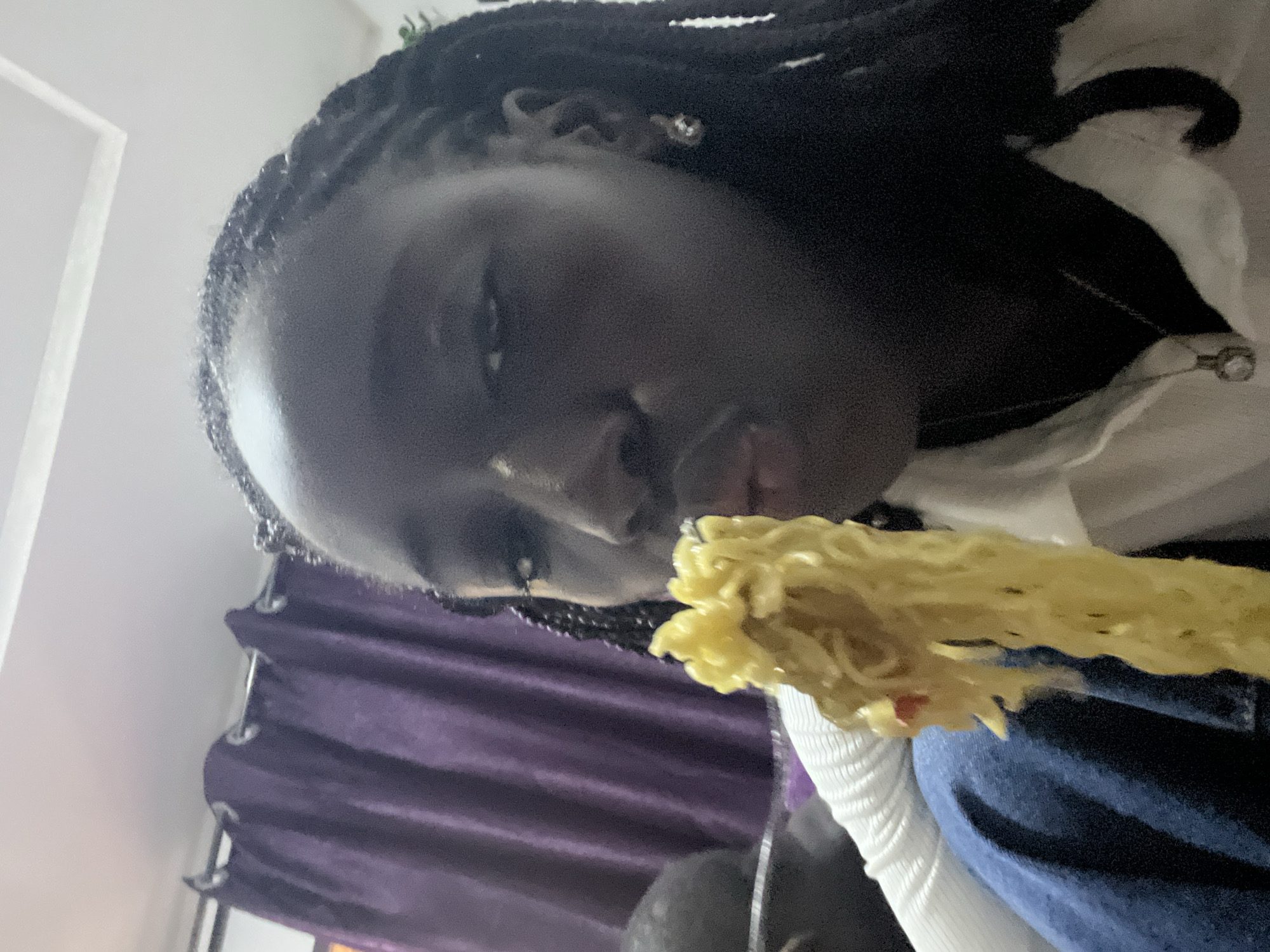 A rotated, close-up image captures a person with dark skin and braided hair, engaged in eating. The person, positioned diagonally across the frame, is holding a substantial portion of yellow instant noodles towards their mouth, making the food a central focus of the shot. They are wearing a white ribbed long-sleeved top, and a silver necklace with a small pendant is visible around their neck. A small earring can be seen on their right ear.
The background is softly blurred but reveals elements of an indoor setting. To the left, a bright white wall or ceiling is visible, while the lower portion features pleated purple curtains with silver grommets, suggesting a window or room divider. The person's attire and the casual setting imply a relaxed home environment. The lighting is consistent with indoor conditions, and the time of day is not explicitly discernable. The person's expression appears neutral and focused on the meal. No visible text is present in the image. The context for the location is Abuja, Nigeria.