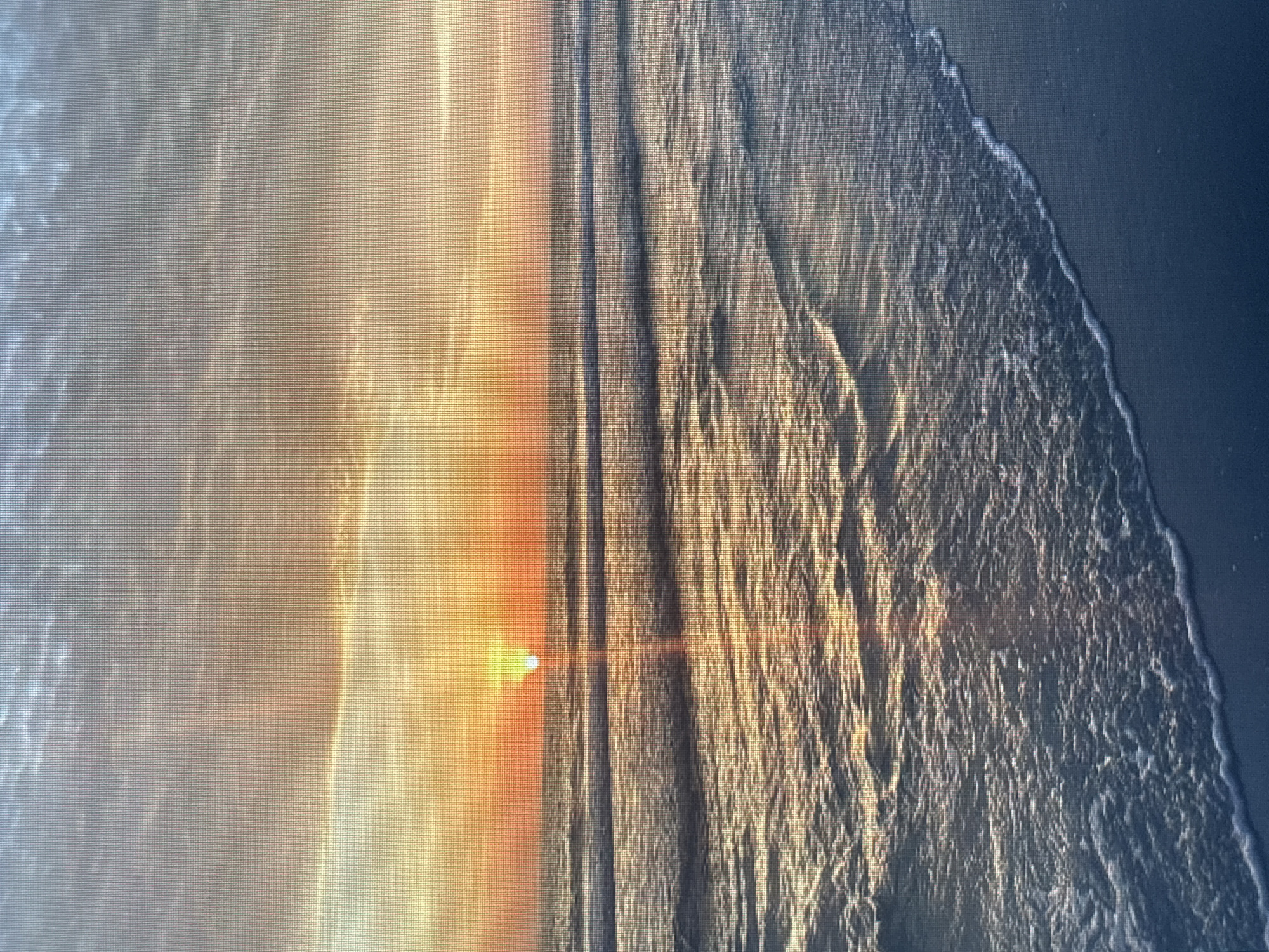 The image depicts a sunset over the Black Sea, likely captured near Varna, Bulgaria. The sun is low on the horizon, casting a golden glow across the water. Waves are visible, and a sandy beach is discernible. The scene conveys a sense of tranquility.