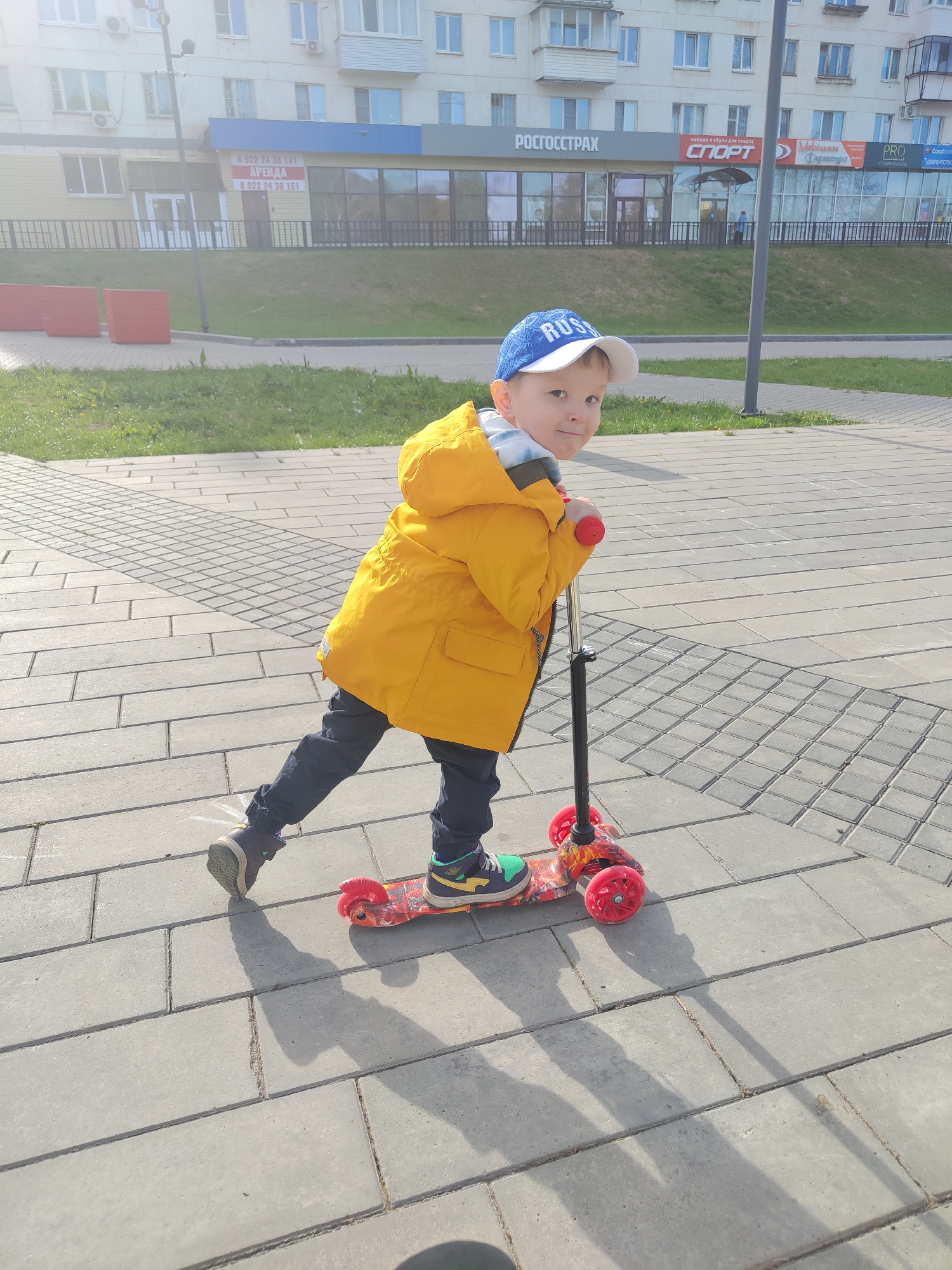 The image shows a child riding a scooter on a paved area in Chaikovsky, Russia. The child wears a yellow jacket and a blue and white cap. The scooter is red and black. In the background, there is a building with several signs in Russian, including "ROSGOSSTRAKH" and "SPORT". There is also an advertisement for rentals (ARENDA) with phone numbers. The environment appears to be an urban area with a mix of commercial and residential buildings.