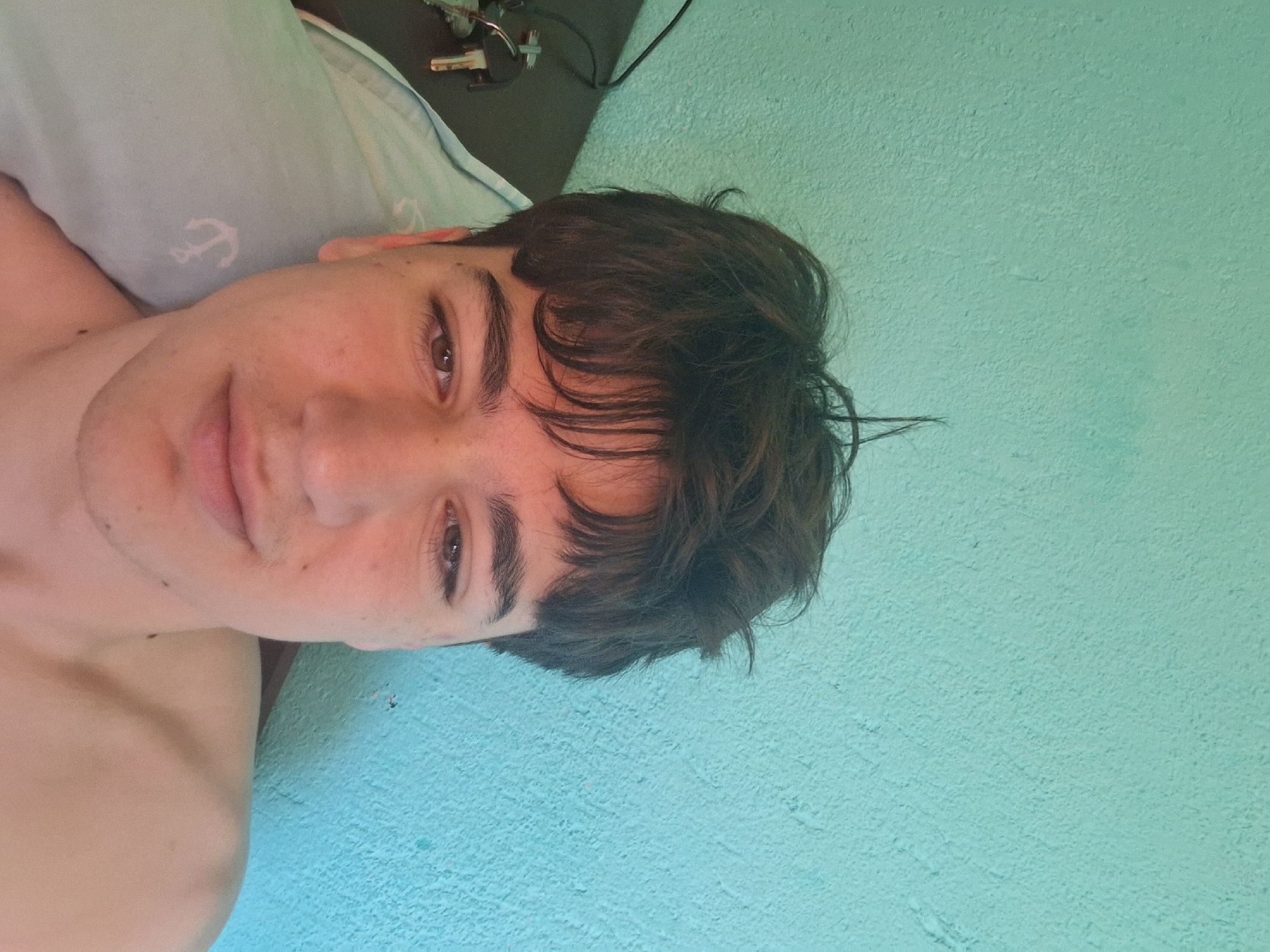 A selfie of a light-skinned young man with brown hair, lying down on a light blue pillow with an anchor design. He is facing the camera, with a neutral expression. Part of his bare shoulder is visible. In the background, there's a turquoise wall and some objects on a dark surface, including keys. The location is not identifiable as Stara Zagora, Bulgaria, without additional information.