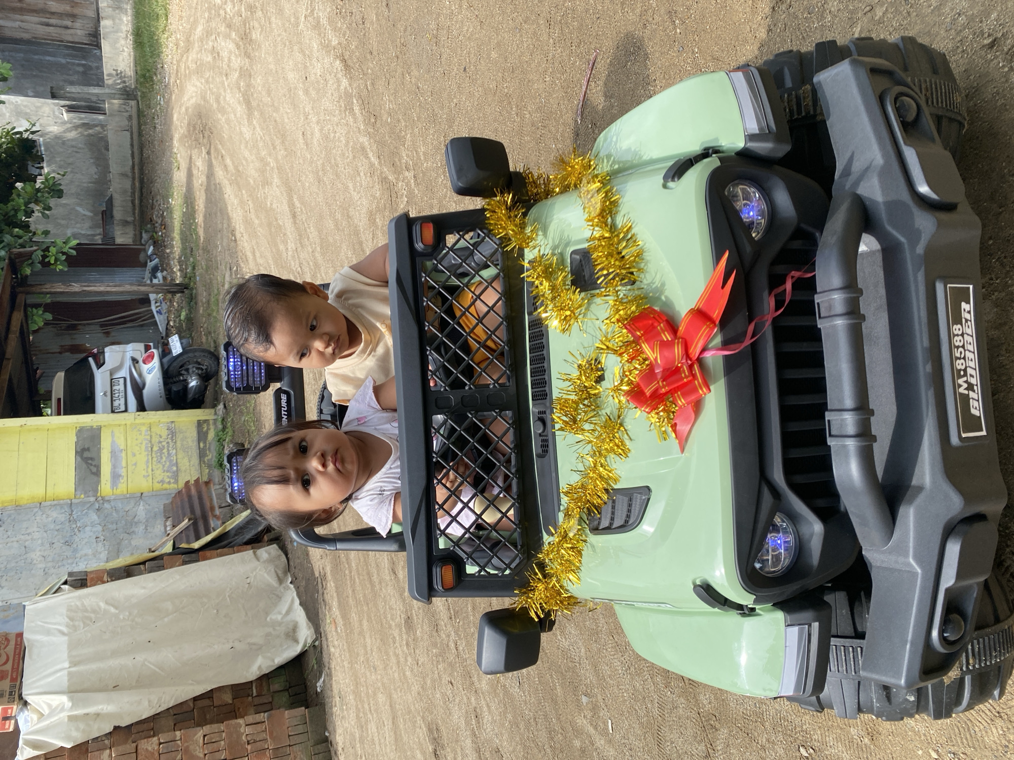 Two young children are sitting in a toy jeep decorated with gold tinsel and a red bow. The jeep is green with a black front grille. The vehicle model is labeled "M-8588 BLOBBER". The children are looking towards the camera. The background shows a yellow building, corrugated metal structures, and brick piles. Other vehicles are partially visible. The ground is paved with a light brown surface. The location is Lhokseumawe, Indonesia.