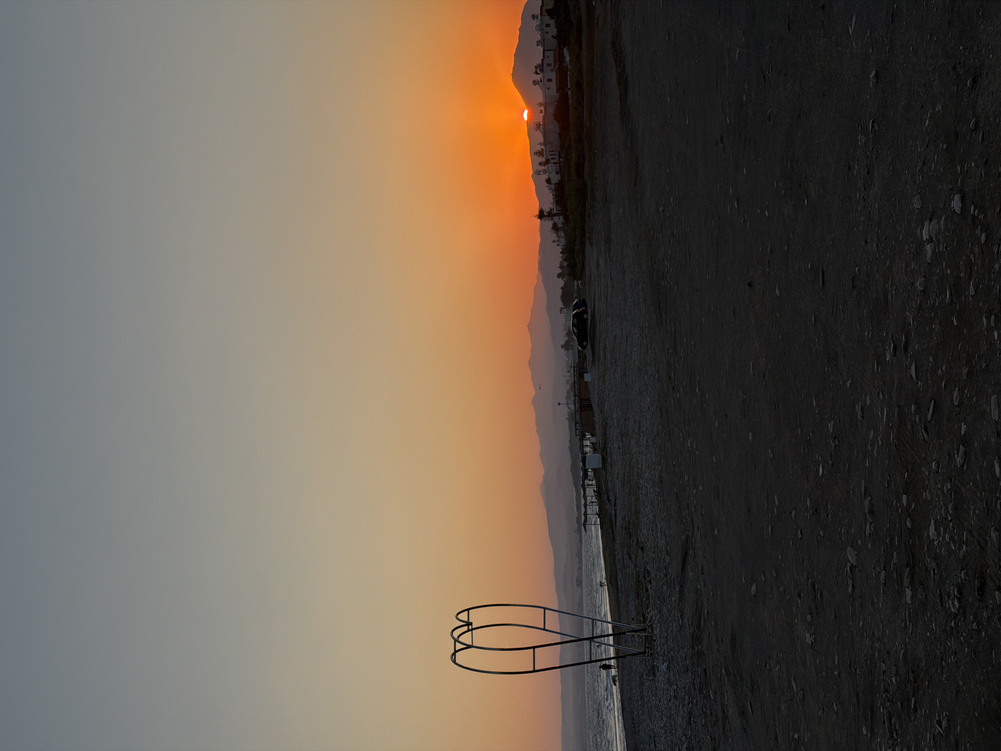 The image depicts a sunset over the beach in Meneou, Cyprus. The sky transitions from a pale blue at the top to a vibrant orange near the horizon, where the sun is setting behind a mountain range. In the foreground, the dark sand of the beach is dotted with small stones. To the left, a distinctive metal sculpture with a heart-like shape stands near the water's edge. A single person and a dog are visible walking on the wet sand closer to the sea. Further down the beach, several beach umbrellas are set up. In the middle ground, a dark-colored car is parked on the sand. Buildings with white roofs are visible on the slopes of the mountains. The sea is calm, with gentle waves breaking on the shore.