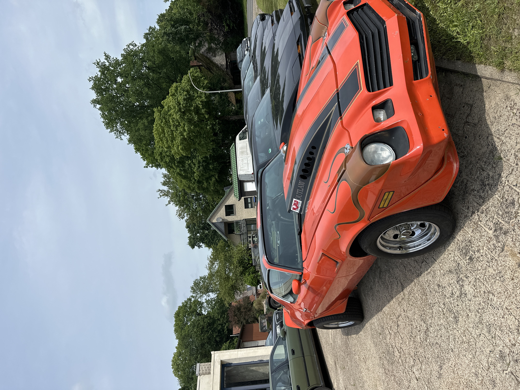 The image shows a line of cars parked outdoors on a paved surface next to a grassy area. In the foreground, an orange American muscle car with black racing stripes on the hood and a prominent hood scoop is parked. The car has custom flame graphics on the side. Behind it, a series of black cars, which appear to be Pontiac Firebirds based on their shape and pop-up headlights, are parked in a row. Further back, a green sedan is visible on the left, and a military-style vehicle is parked near a building on the right. Several houses and trees are visible in the background, under a cloudy sky. The scene is located in Kutenholz, Germany, as indicated by the presence of a white van with "Kutenholz" written on it, partially visible behind the orange muscle car. There are no people visible in the image.